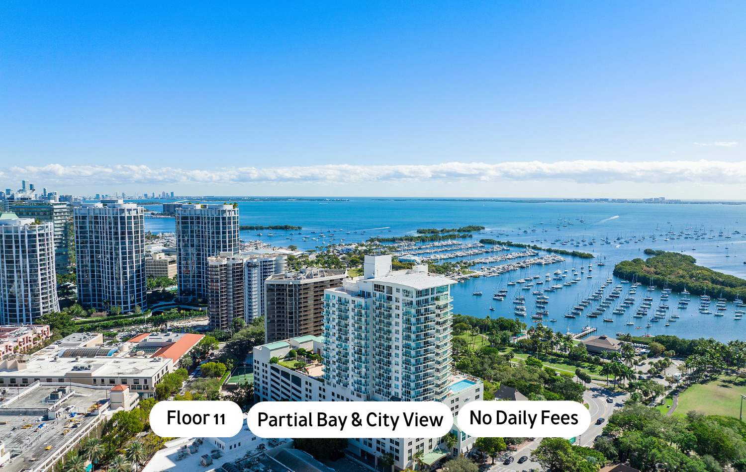 Panoramic aerial view showcasing a vibrant city skyline with numerous high-rise buildings, a wide expanse of blue bay dotted with boats and marinas, and a clear sky with scattered clouds.