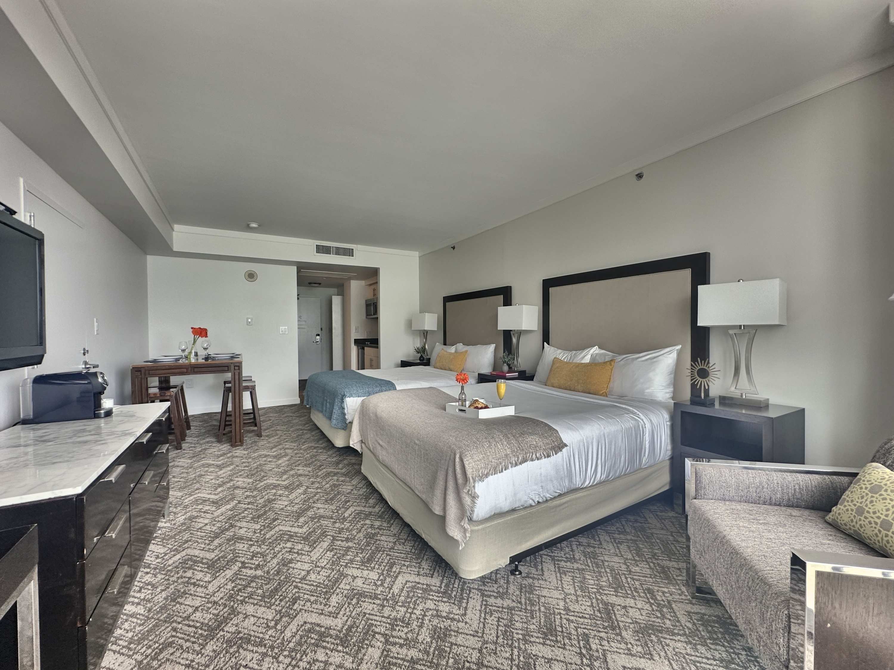 An expansive view of the studio apartment, showcasing two queen beds with elegant headboards, a dining table with stools, a modern dresser with a TV, and a cozy armchair, all on patterned carpet.