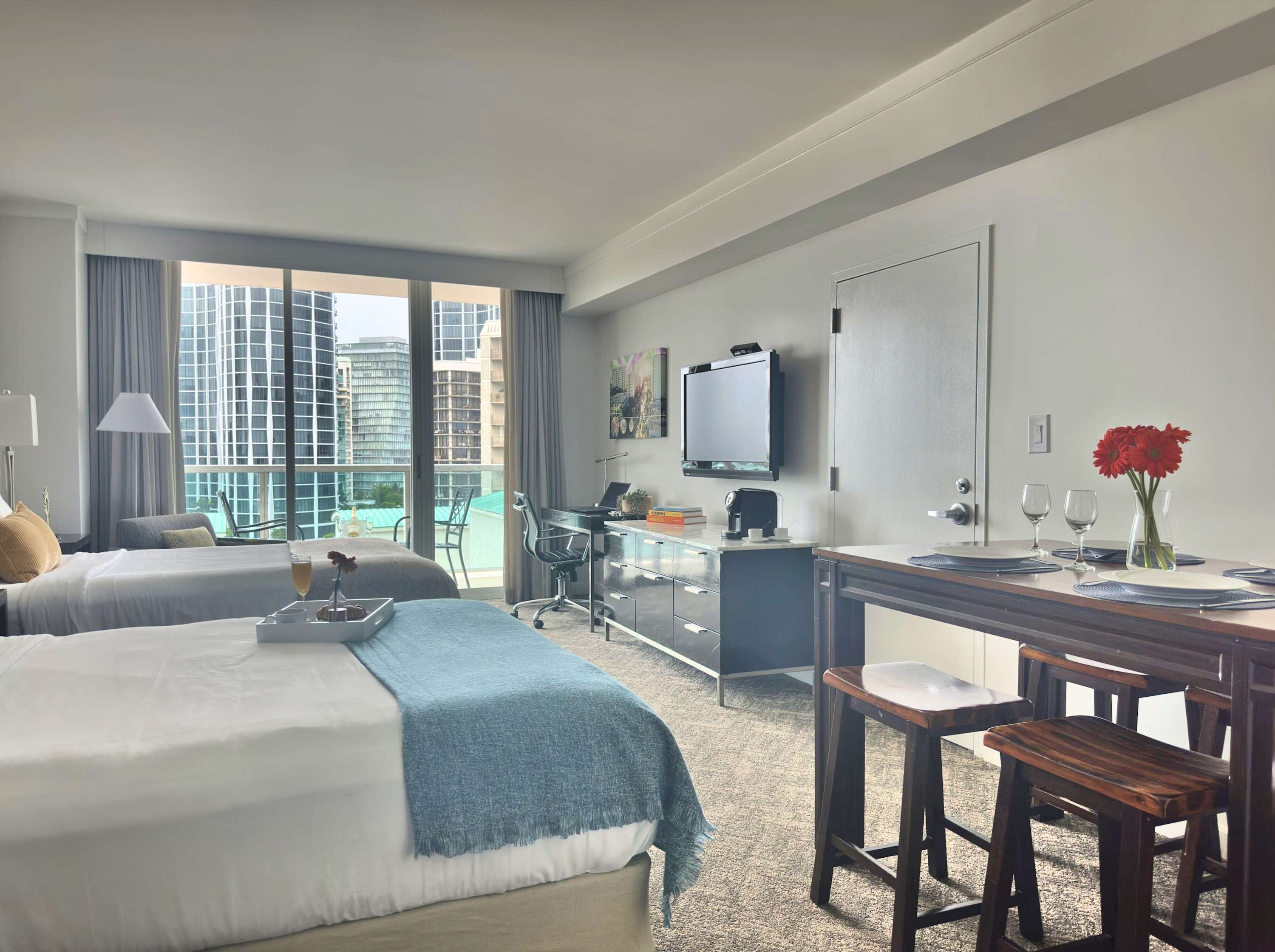 A spacious studio room with two beds, a high dining table with stools, and a dedicated workspace with a desk and chair, all facing a large window offering views of surrounding city buildings.