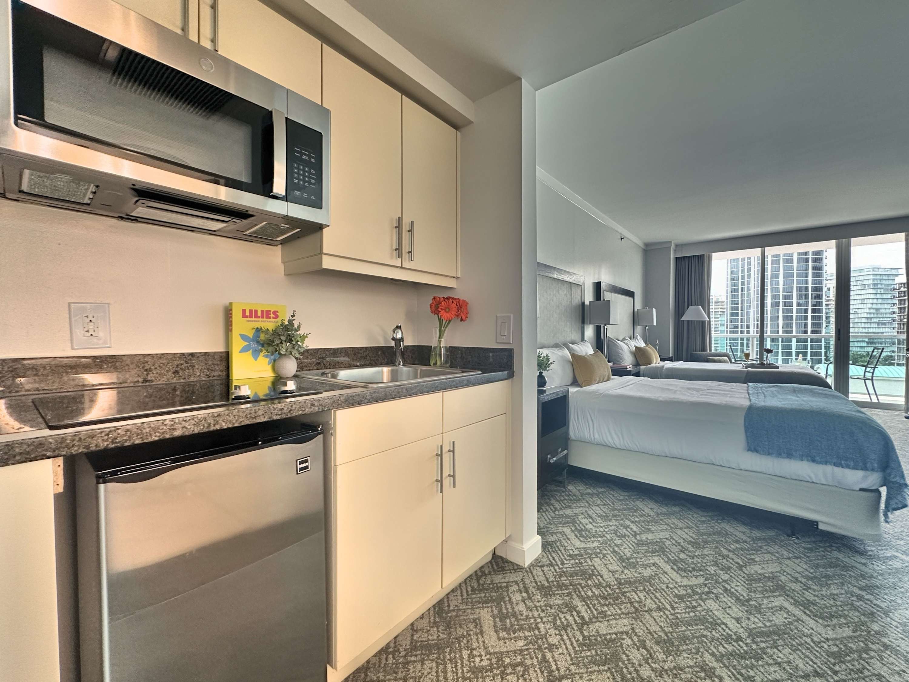A well-appointed kitchenette with cream cabinets, a stainless steel microwave, and a mini-fridge, seamlessly integrated into the studio layout, offering a glimpse into the sleeping area with two beds.