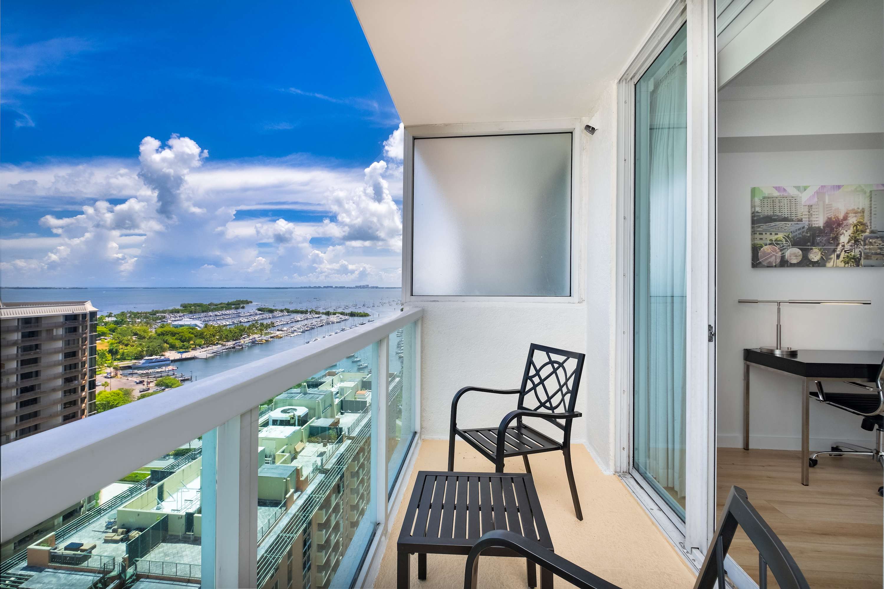 A private balcony featuring black metal outdoor furniture, including two chairs and a small table, offering a stunning partial view of the bay with boats and distant city buildings under a bright blue sky.