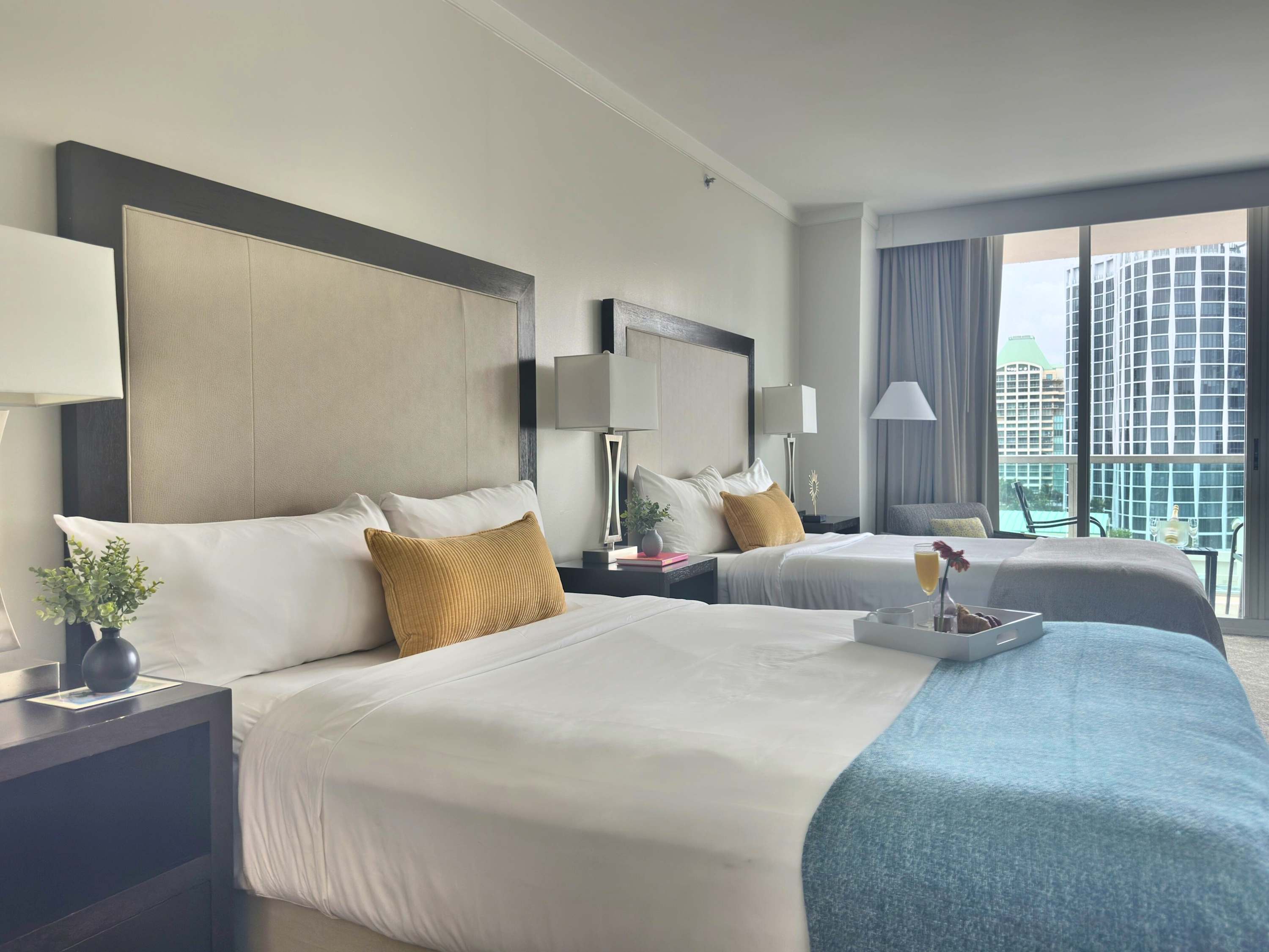 A comfortable queen-sized bed with crisp white linens, a light blue throw, and a yellow accent pillow, featuring a breakfast tray with juice and pastries, positioned next to a nightstand with a lamp.