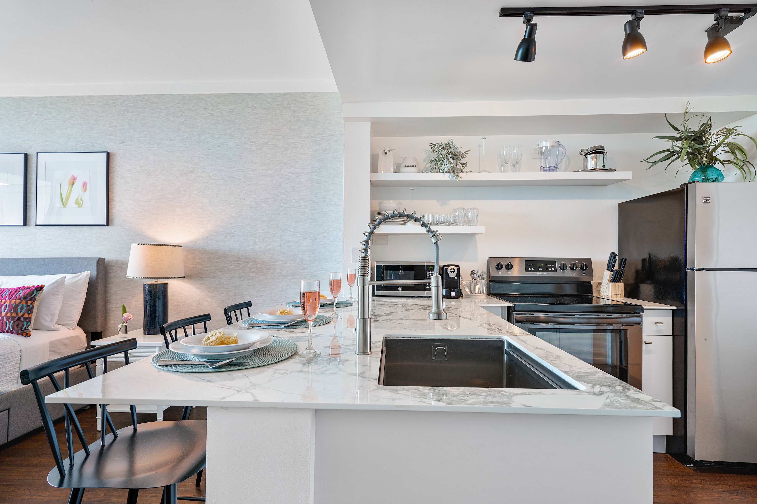 Modern kitchen island with a deep stainless steel sink and a flexible faucet, surrounded by three black bar stools, and open shelving above a black refrigerator and electric stove.