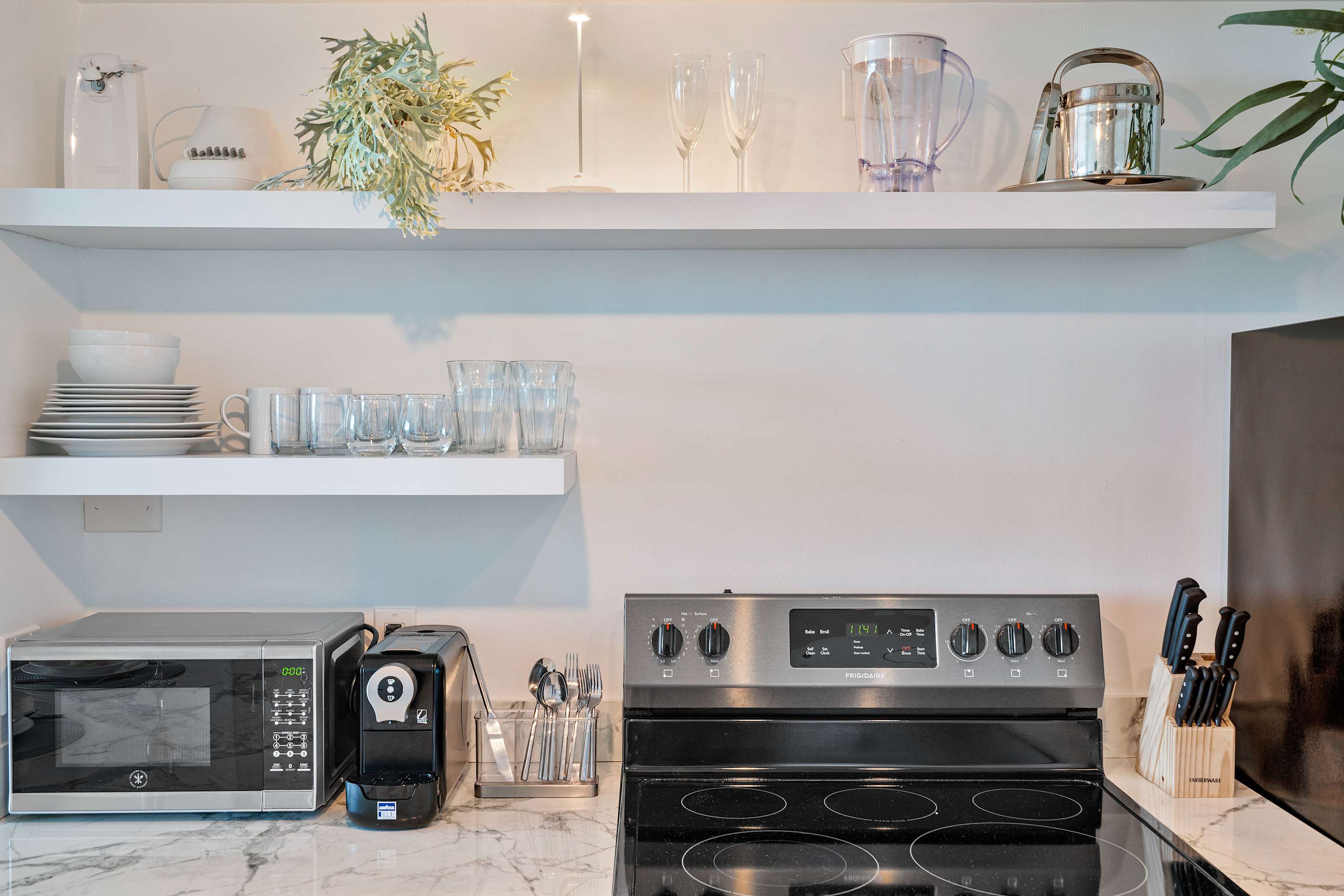 Detailed view of a contemporary kitchen counter featuring a black electric stove, microwave, coffee maker, and open shelves displaying various kitchenware and decorative plants.
