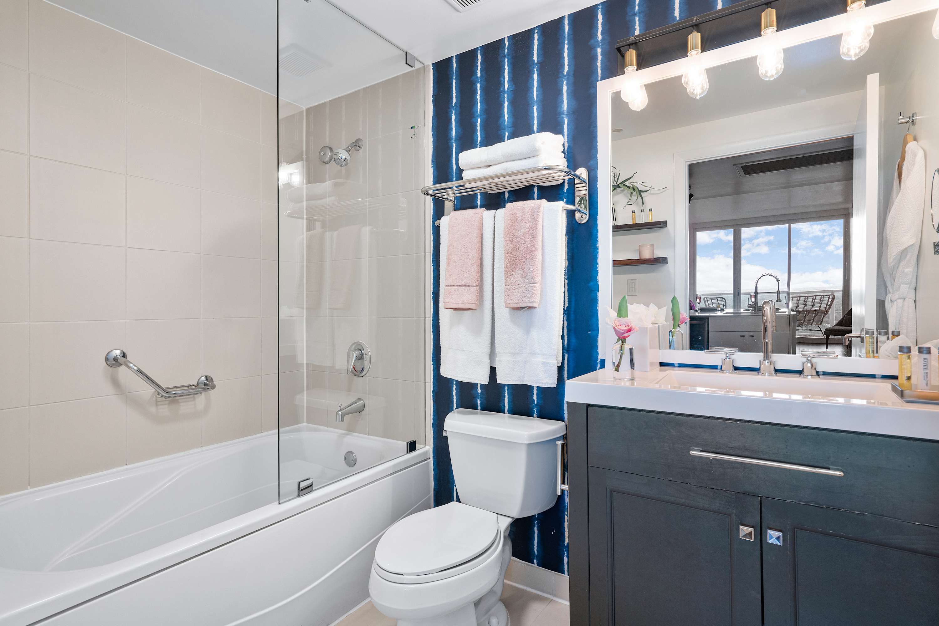 A contemporary bathroom with a white toilet, a bathtub with a glass shower screen, and a dark vanity, featuring a striking blue and white accent wall and light-colored tiled walls.