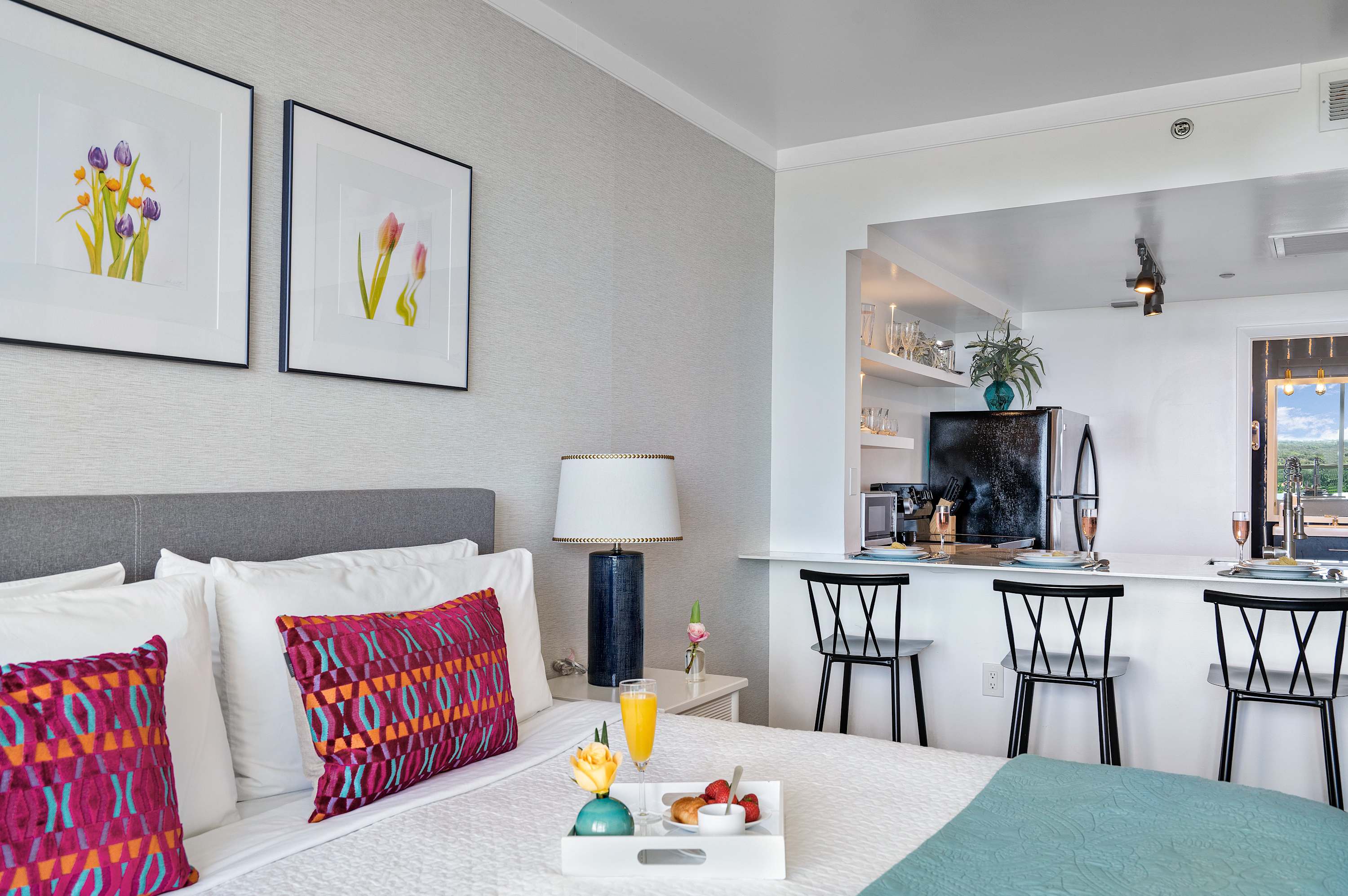 A bright studio bedroom with a white bed, colorful pillows, and a breakfast tray, featuring a compact kitchenette with a black fridge and bar seating in the background.