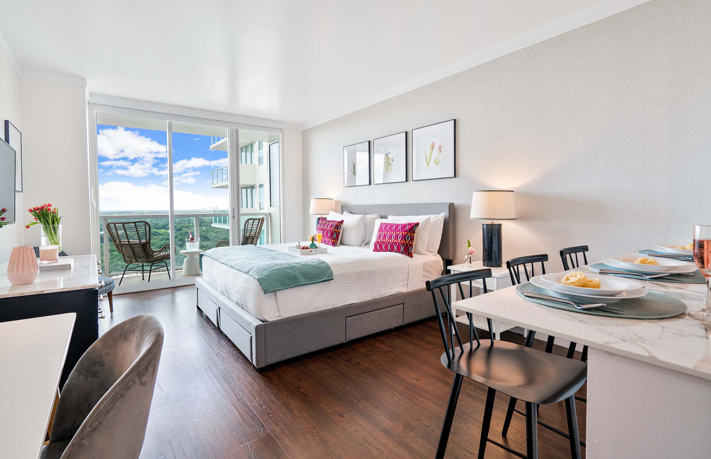 Wide view of a bright studio apartment featuring a queen bed, a dining area with a marble table and black chairs, and a small desk, all with hardwood floors and a balcony view.