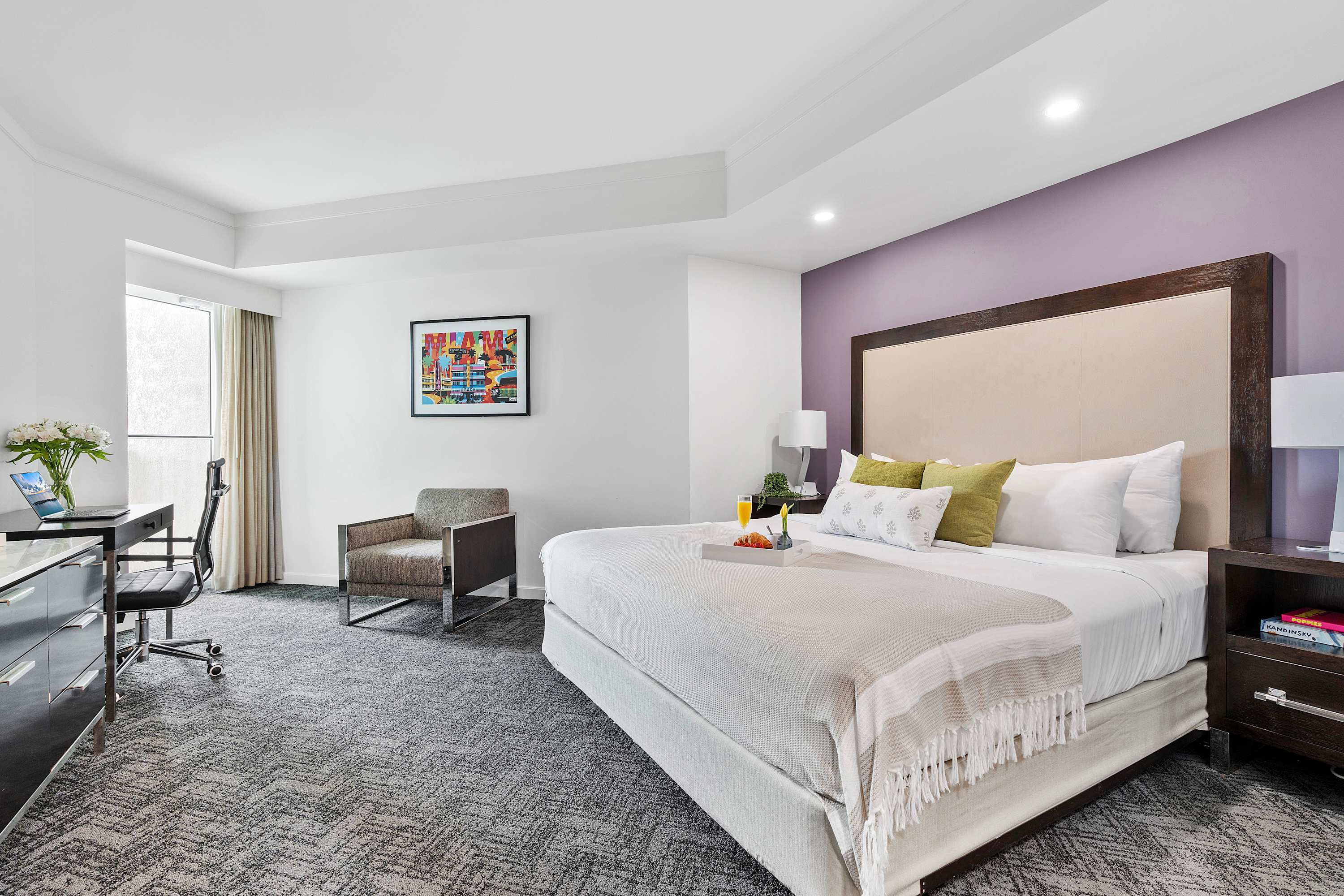 Modern bedroom with a king-sized bed featuring a tall upholstered headboard against a light purple accent wall, grey patterned carpet, a desk with a chair, and a separate armchair. A breakfast tray with fruit and juice is on the bed.