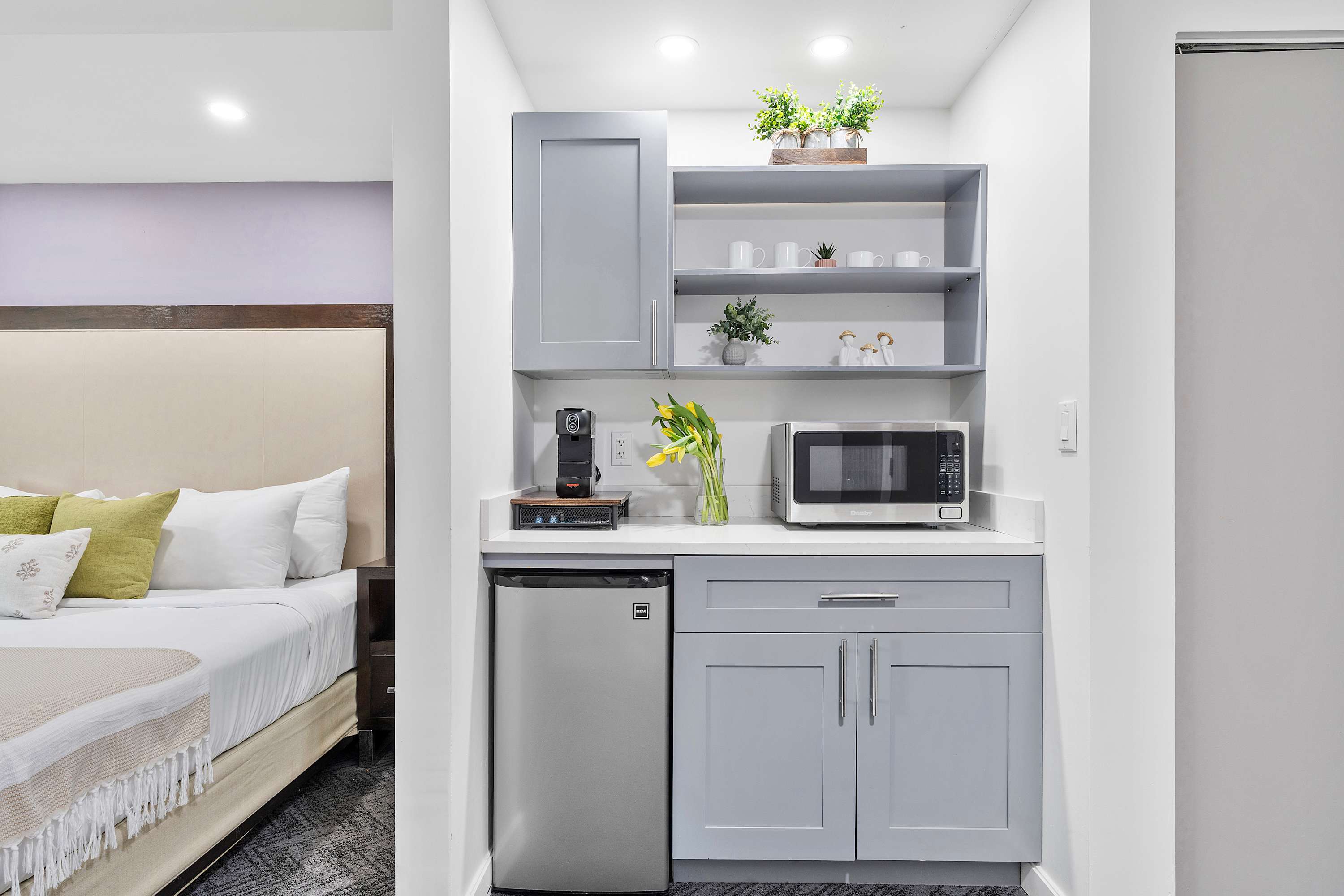 Compact kitchenette area with light grey shaker-style cabinets, white countertops, a stainless steel mini-fridge, microwave, and coffee maker. Open shelving displays mugs and decorative plants.