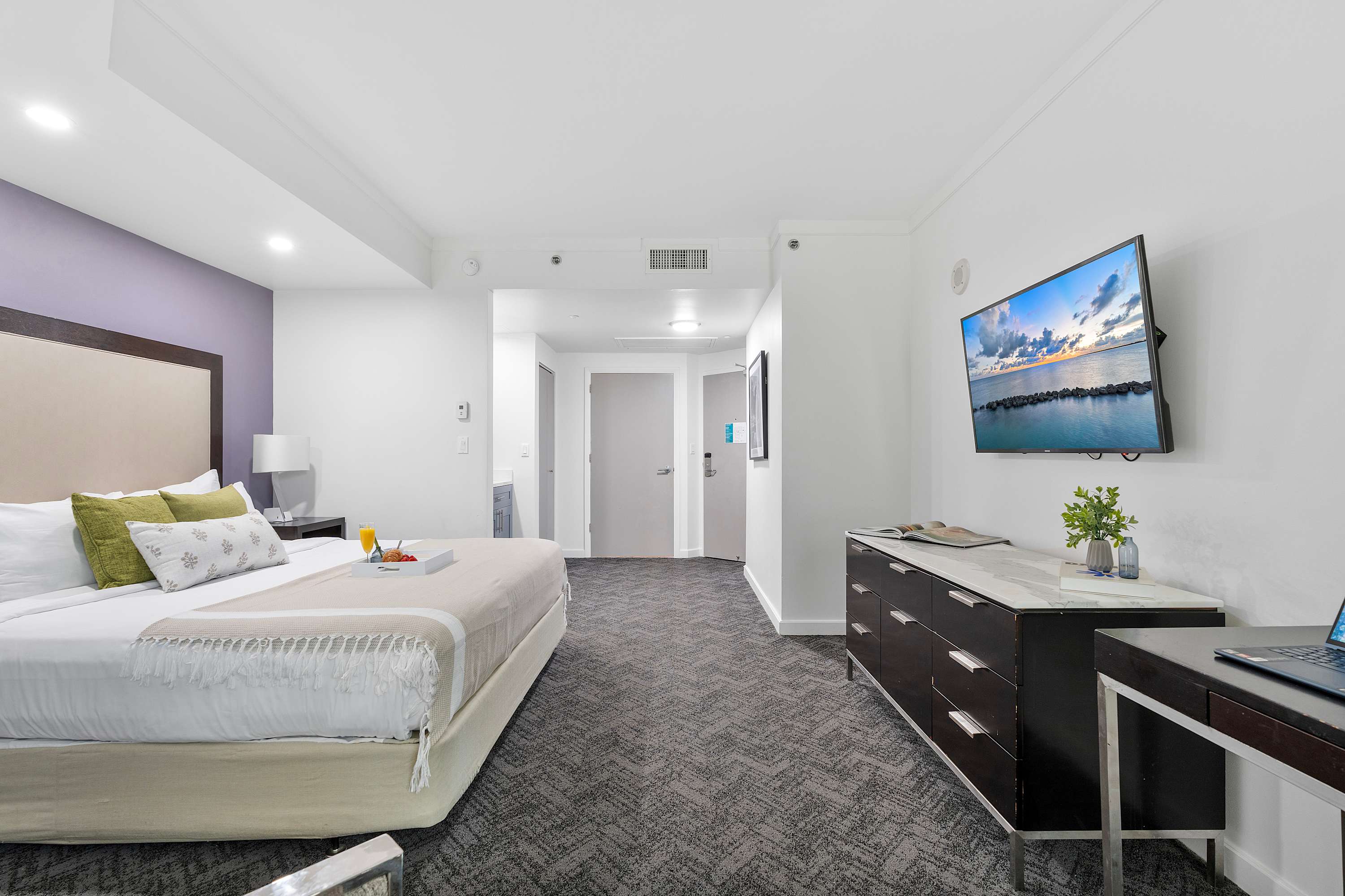 Wide view of the modern bedroom, showing the king-sized bed, a long dark wood dresser with a wall-mounted flat-screen TV, and the entrance hallway. The room features grey patterned carpet and white walls.
