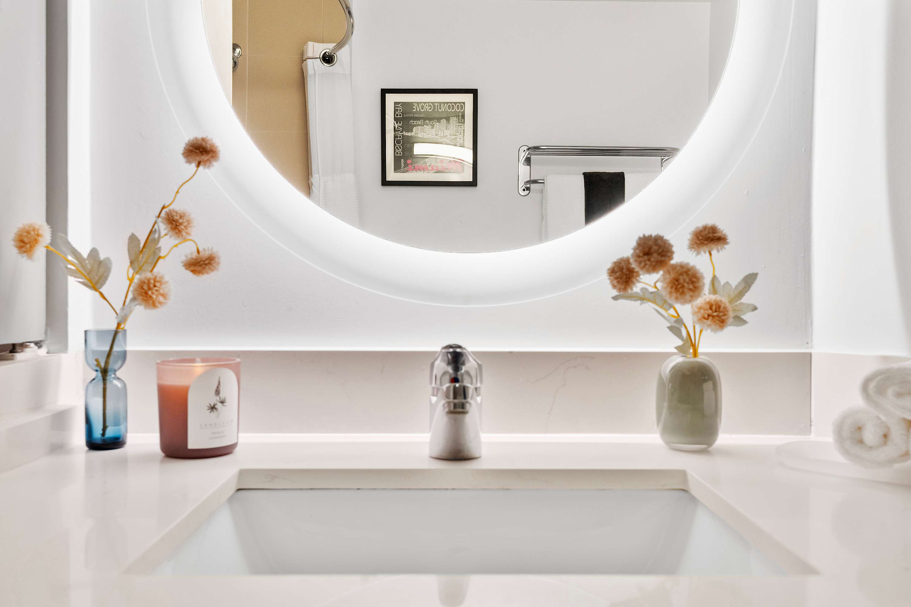 Close-up of a modern bathroom vanity with a white rectangular sink, chrome faucet, and a large circular backlit mirror. Decorative flowers and a candle adorn the white quartz countertop.