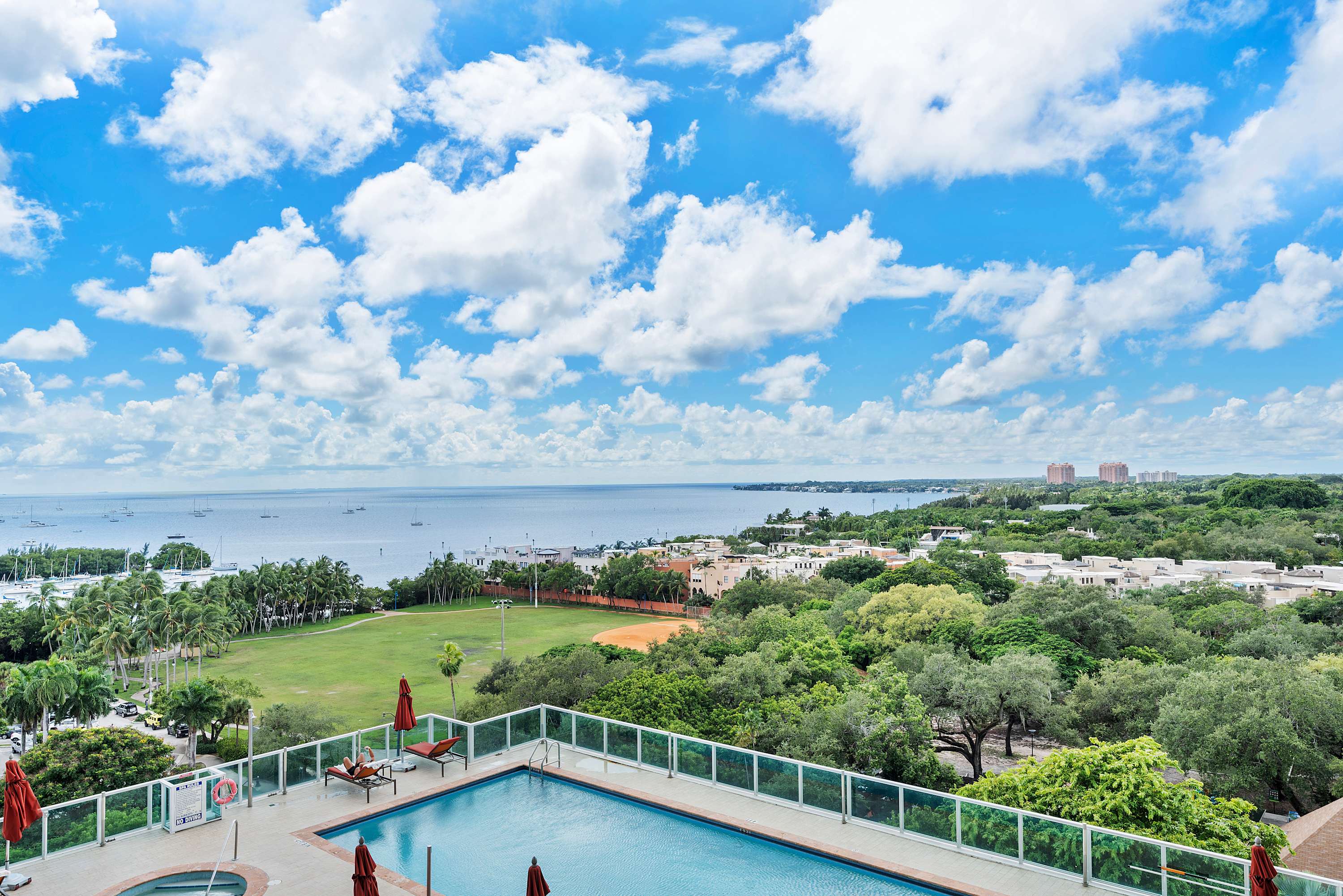 Elevated outdoor swimming pool with blue water, surrounded by lounge chairs and umbrellas, offering a panoramic view of a bay with boats, green spaces, and distant buildings under a blue sky with white clouds.