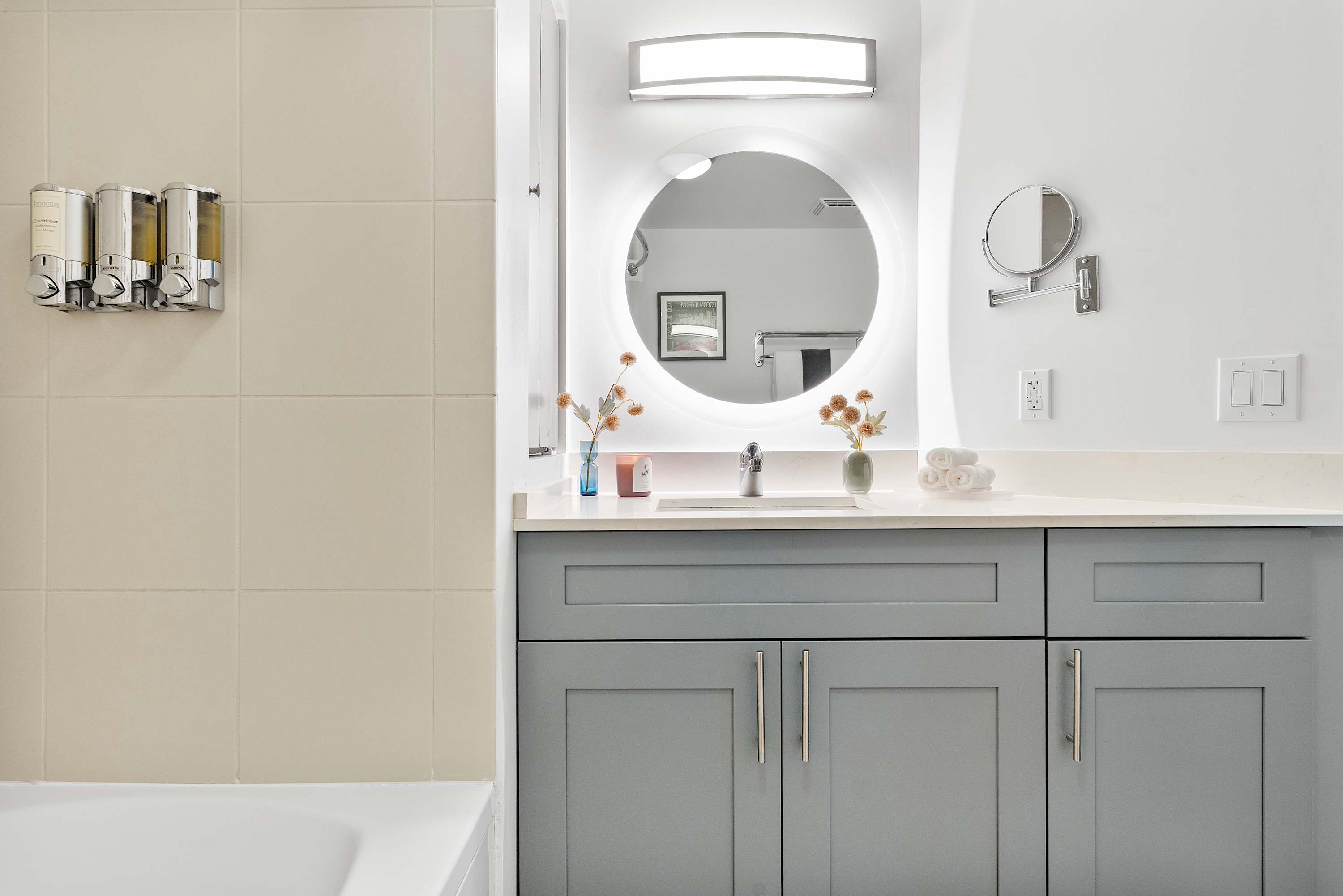 Modern bathroom vanity with grey shaker-style cabinets, a white countertop, and a circular backlit mirror. A wall-mounted magnifying mirror and fresh white towels are also visible.