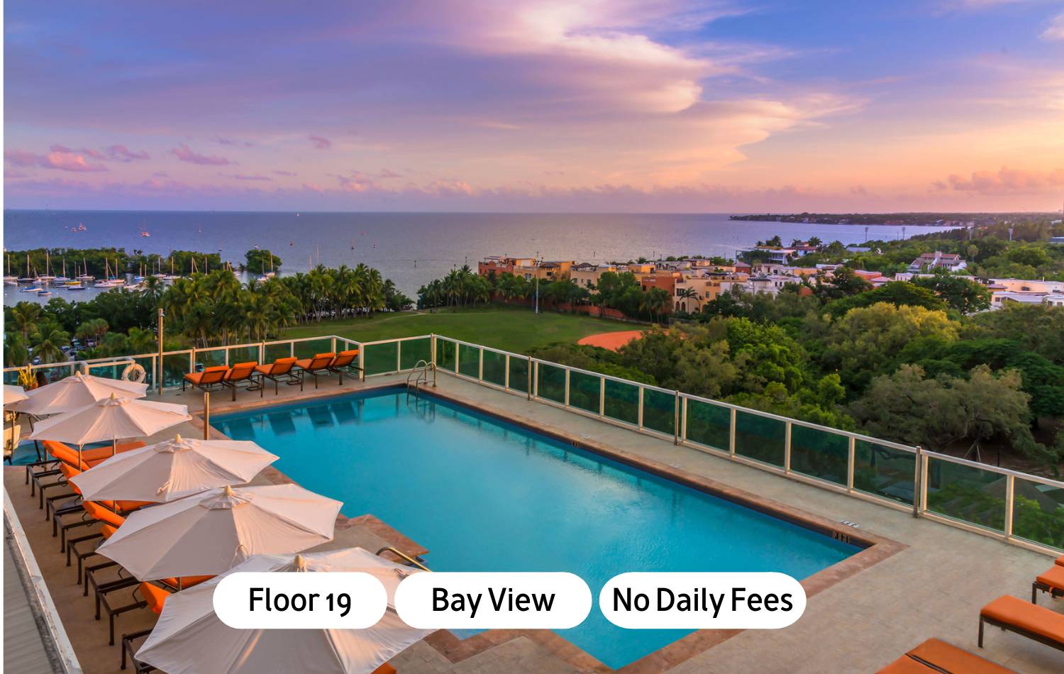Elevated outdoor swimming pool with blue water, surrounded by lounge chairs with orange cushions and white umbrellas, overlooking a bay with boats and a city skyline at sunset.