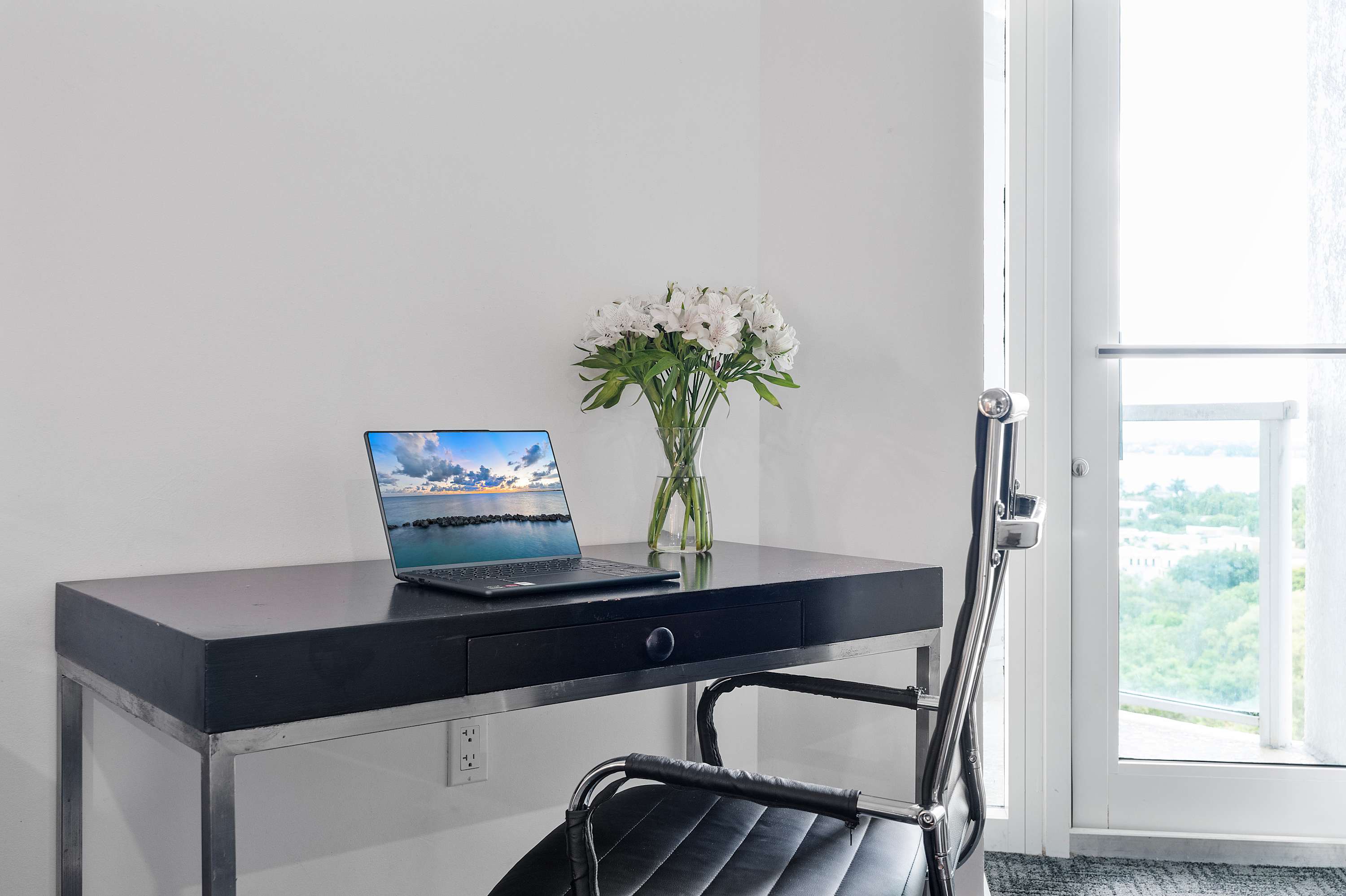 Dedicated workspace featuring a sleek black desk with a silver frame, a black ergonomic office chair, and a laptop displaying a scenic view. A vase of white flowers adds a touch of elegance next to a large window.
