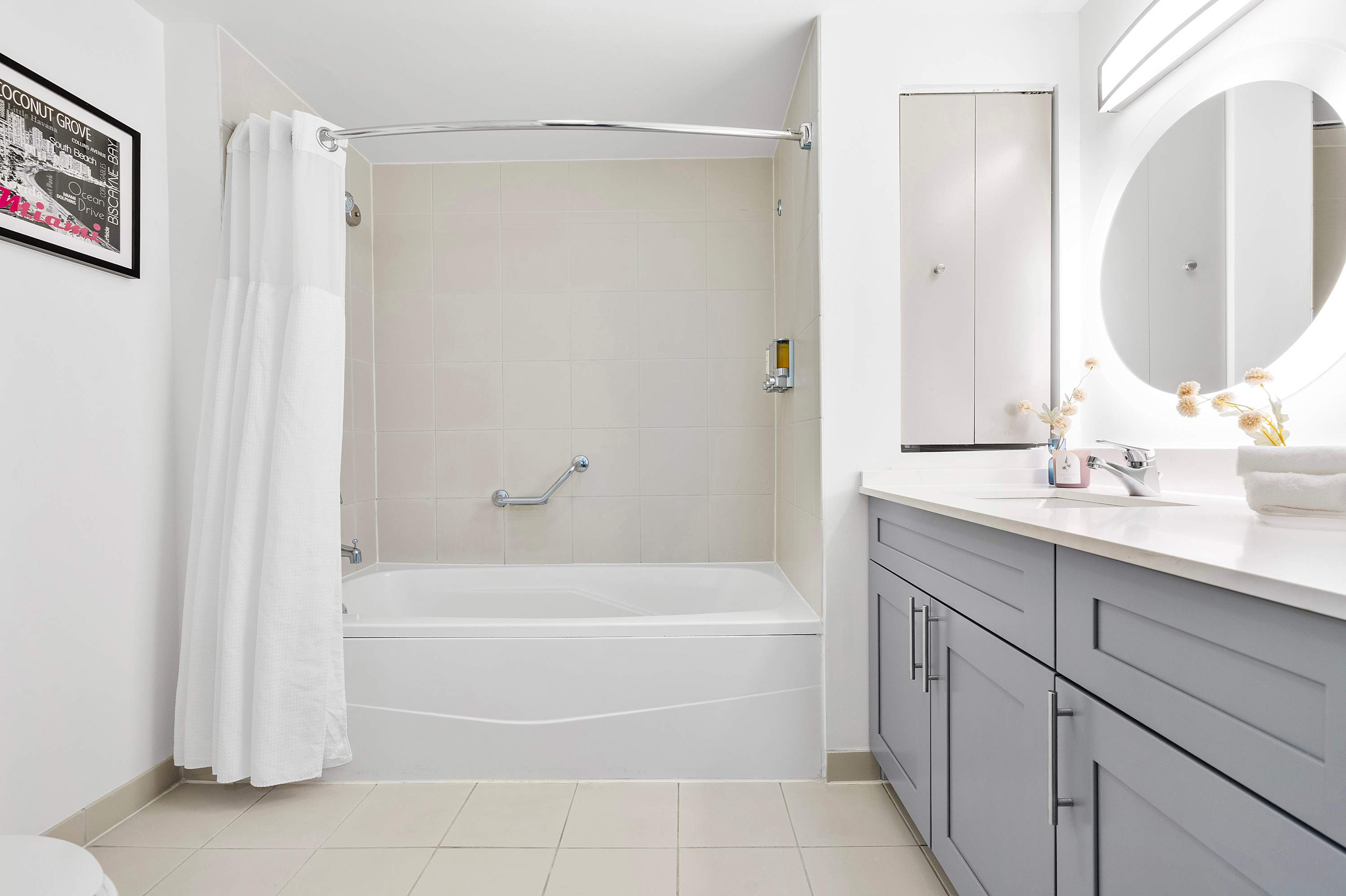 Bright bathroom with a white bathtub and shower combo, light beige tiled walls, and a modern vanity with grey cabinets and a white countertop. A circular backlit mirror hangs above the sink.