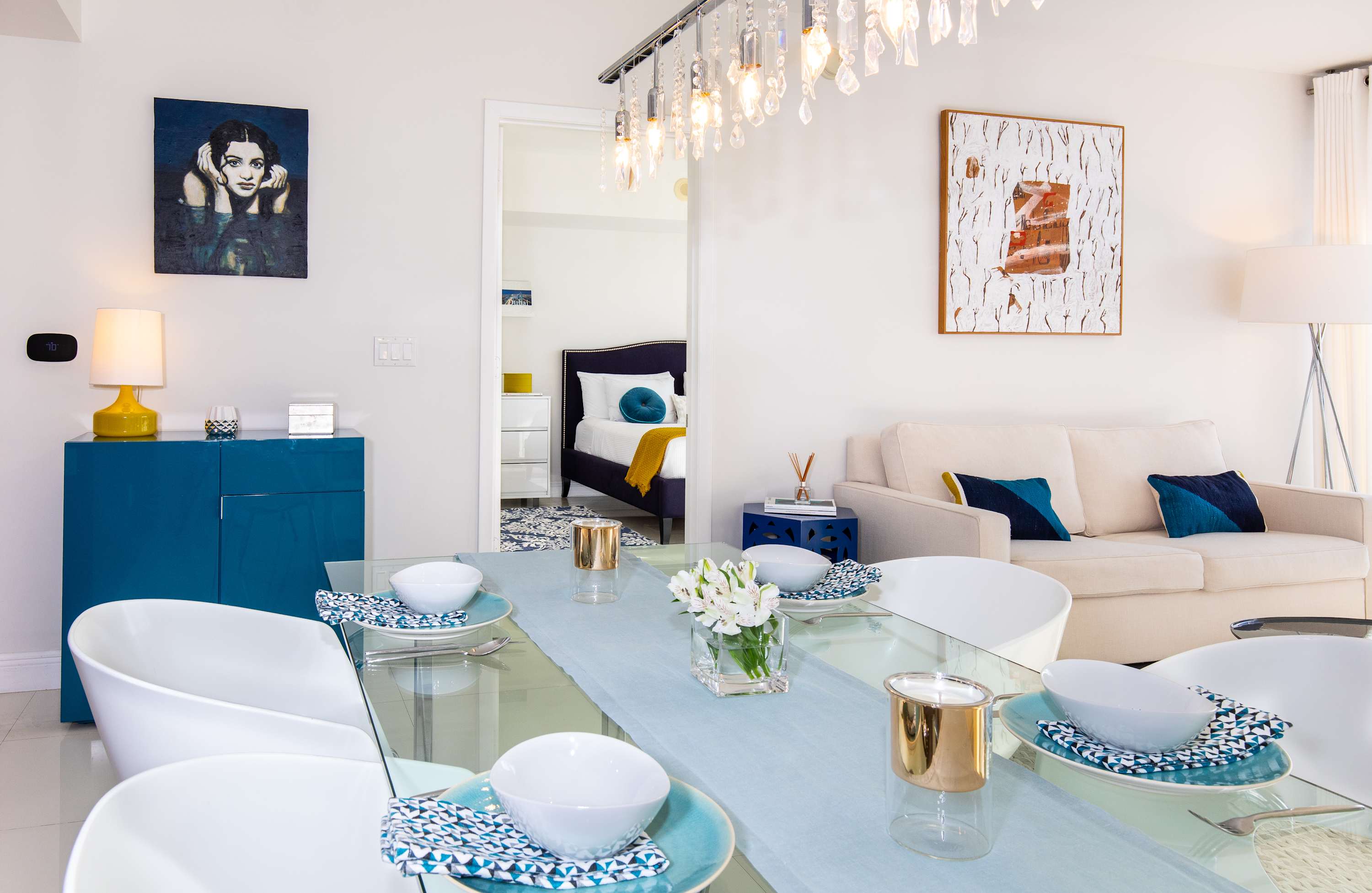 A bright dining area with a glass table set for six, featuring white chairs and a light blue runner. Beyond, a white sofa with blue pillows and contemporary artwork define the adjacent living space.
