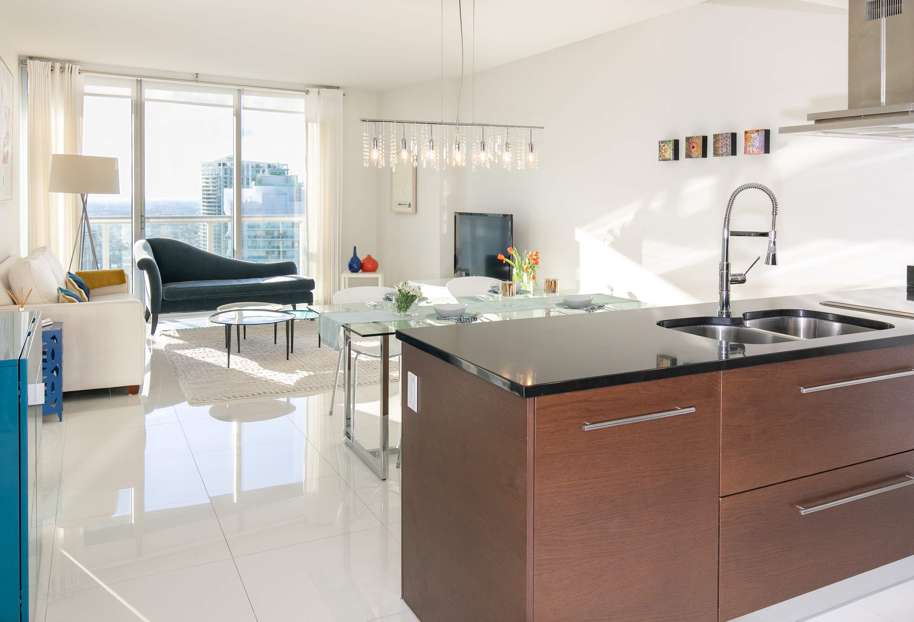 Open concept living space with white tiled floors, a dark blue chaise lounge, a glass dining table, and a modern kitchen island with dark wood cabinets. Large windows offer a city view.