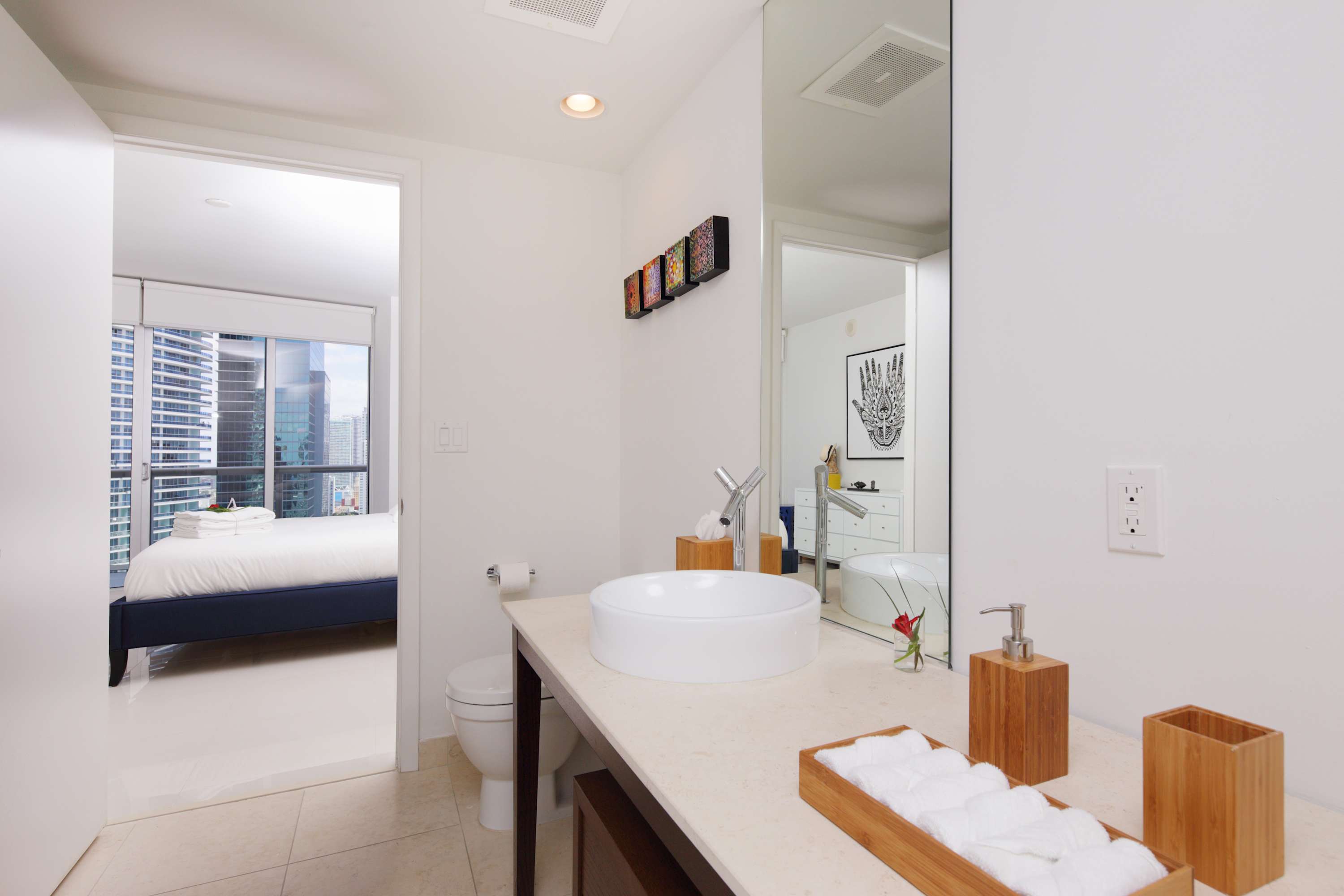 Bathroom with a white vessel sink on a light countertop, a toilet, and bamboo accessories. An open doorway reveals the bedroom with city views in the background.