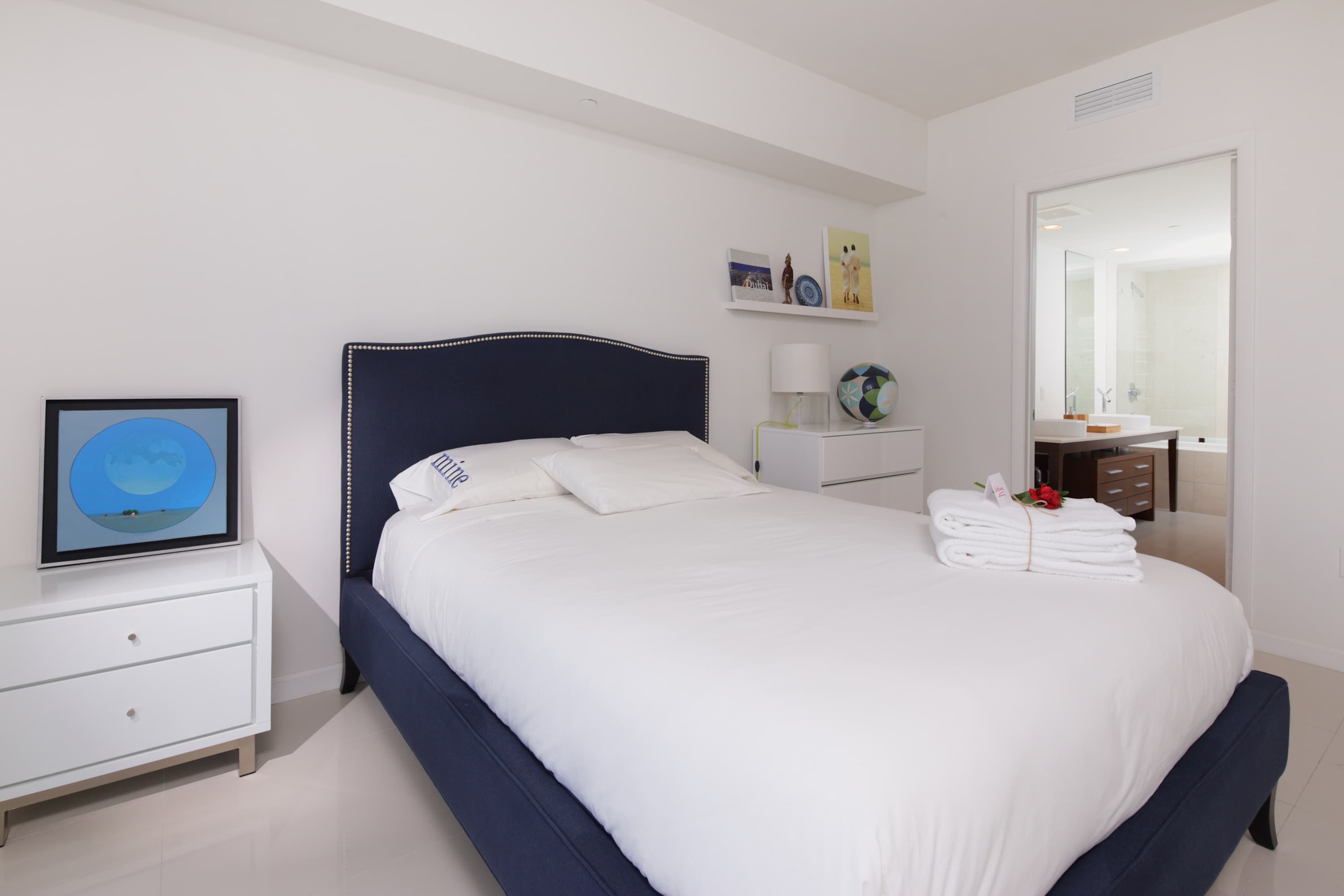 Bedroom with a navy blue upholstered bed frame, white bedding, and a white nightstand with a lamp. A television displaying art is on a white dresser, and a bathroom is visible through an open doorway.