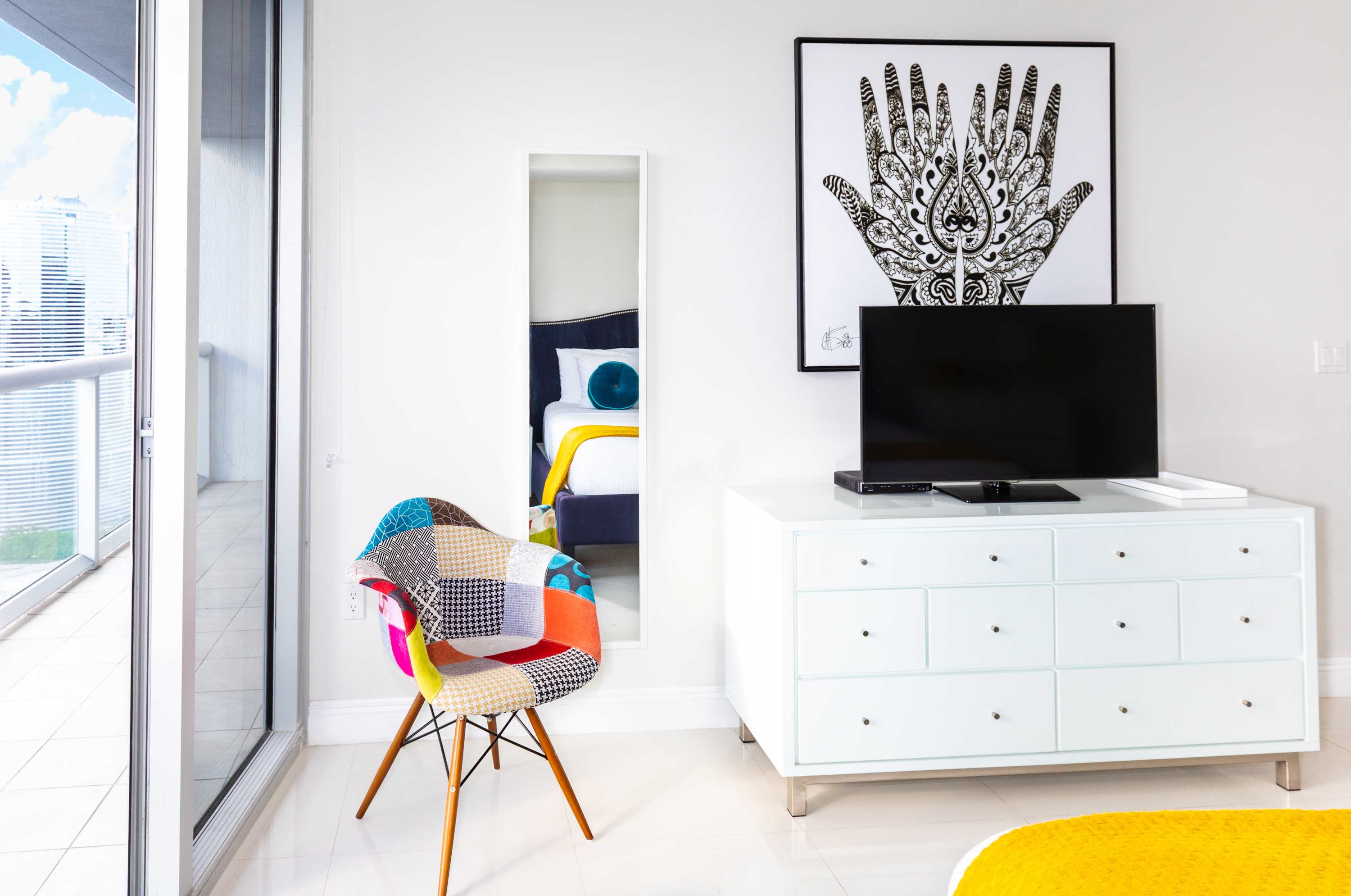 A corner of the bedroom featuring a white dresser with a flat-screen TV, a full-length mirror, and a colorful patchwork chair. Large windows offer a glimpse of the balcony and city outside.
