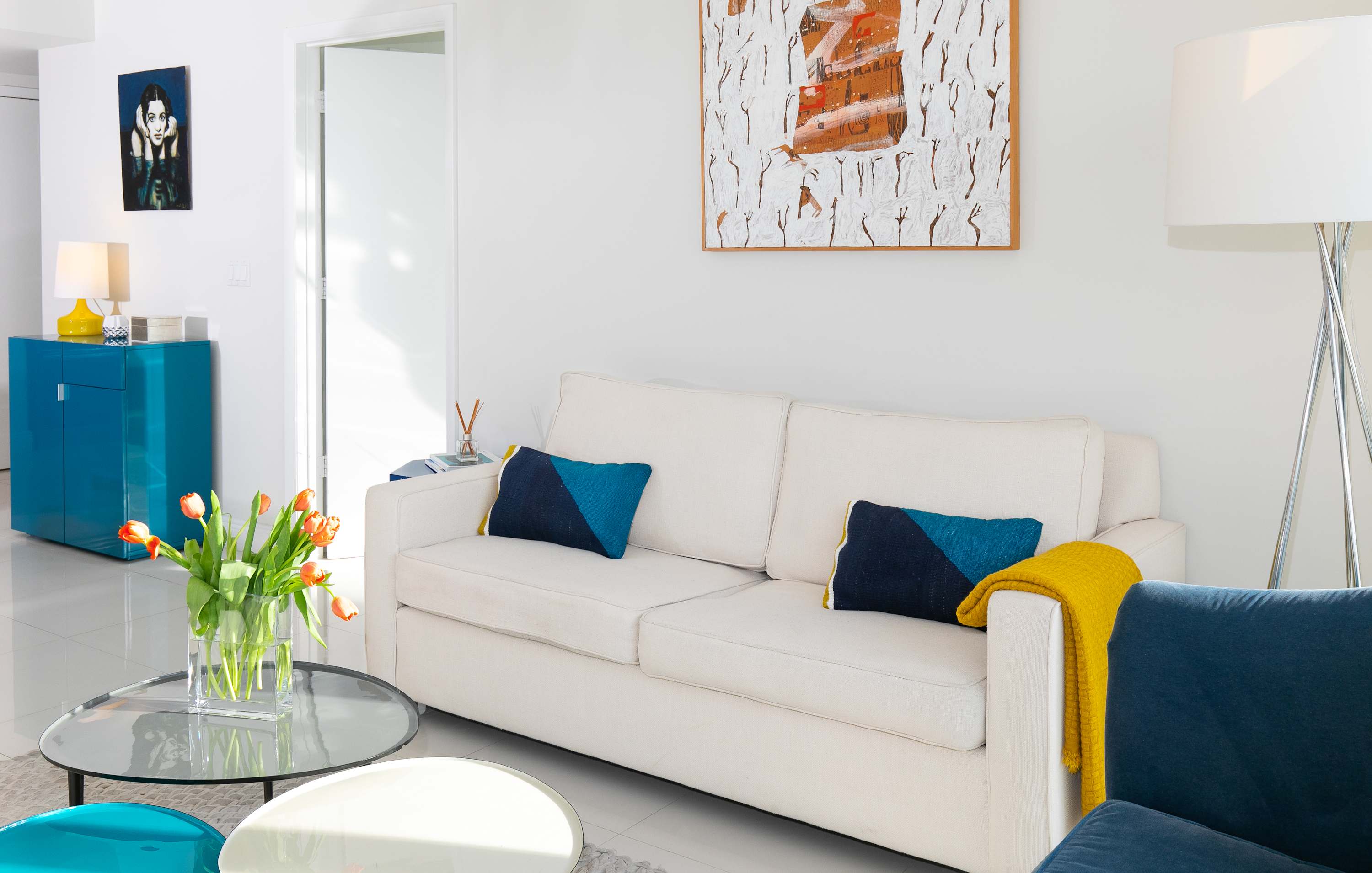 A bright living room with a white sofa adorned with blue and yellow pillows, a blue armchair, and a glass coffee table. Contemporary art hangs on the white walls, and a floor lamp provides additional lighting.
