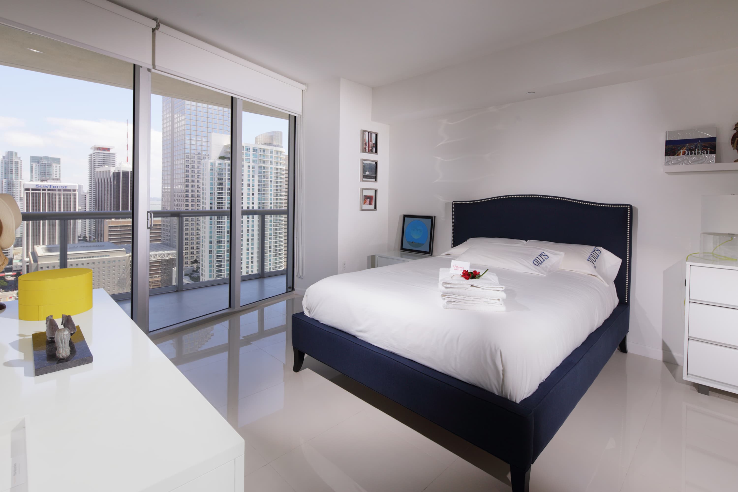 Bright bedroom featuring a navy blue upholstered bed with white linens, white nightstands, and large floor-to-ceiling windows offering expansive city views. A yellow lamp adds a pop of color.