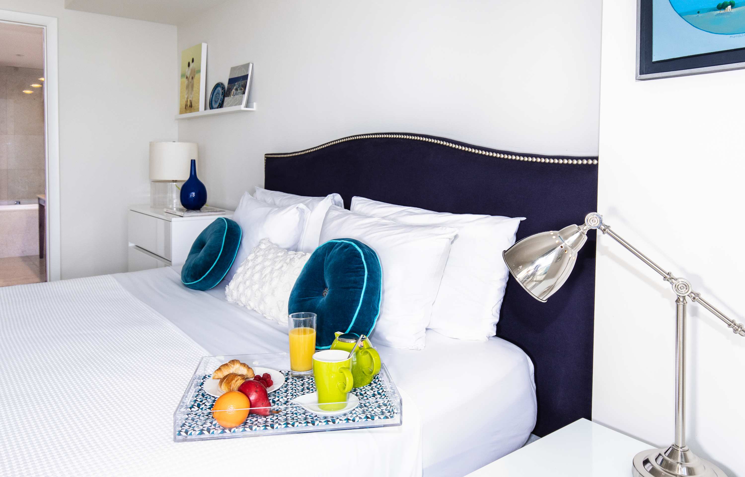Close-up of the navy blue upholstered bed with white bedding and decorative teal pillows. A breakfast tray with juice, coffee, and pastries is on the bed, and a silver desk lamp is on a white nightstand.