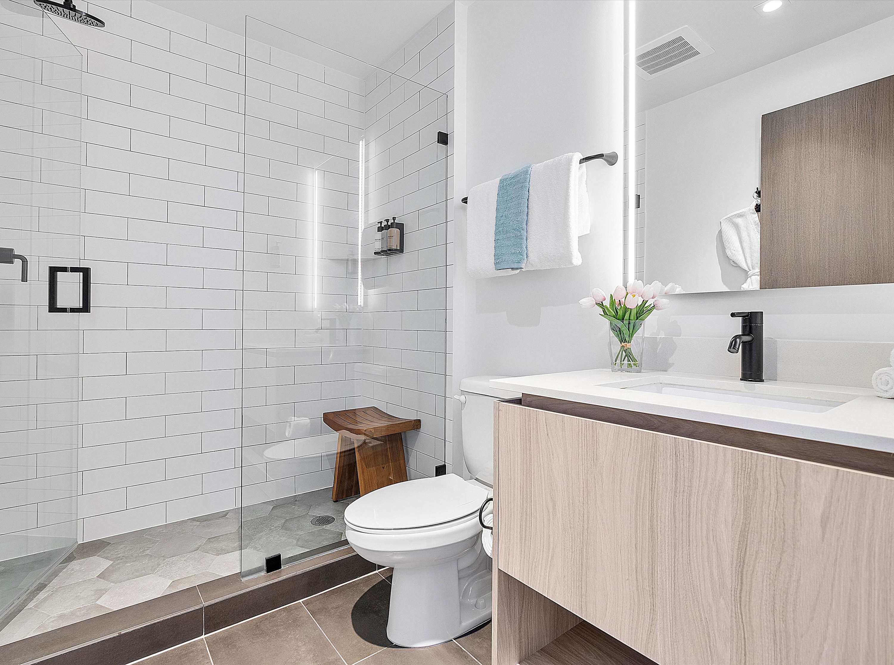 Bright bathroom featuring a light wood vanity with a white countertop, a modern sink, an illuminated mirror, and a walk-in shower with white subway tiles and a glass door.