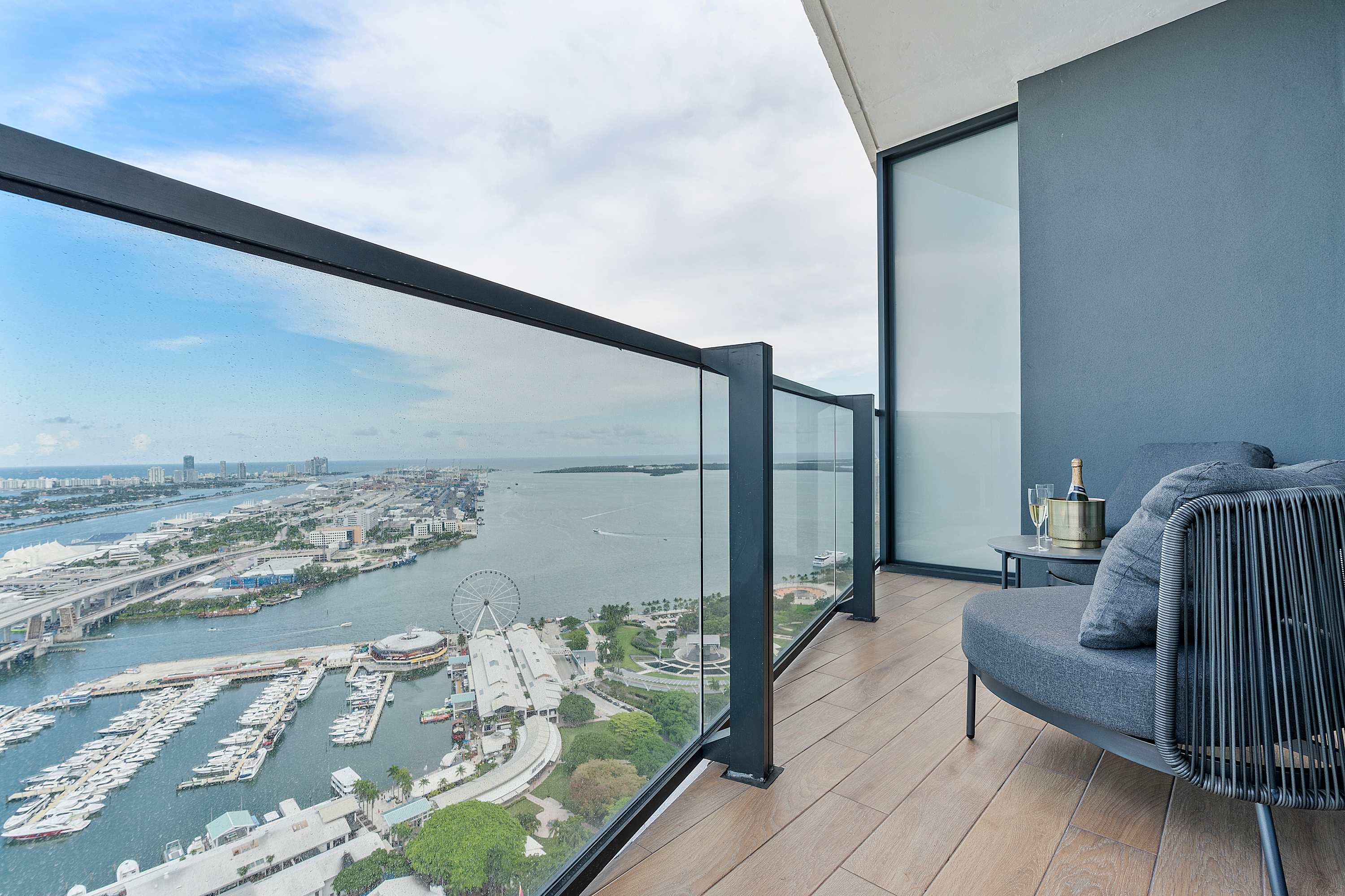 This modern balcony features a sleek glass railing and light wood-look flooring, furnished with a comfortable gray outdoor armchair and a small table holding champagne, offering expansive views of a vibrant cityscape and a bay with boats and a Ferris wheel.