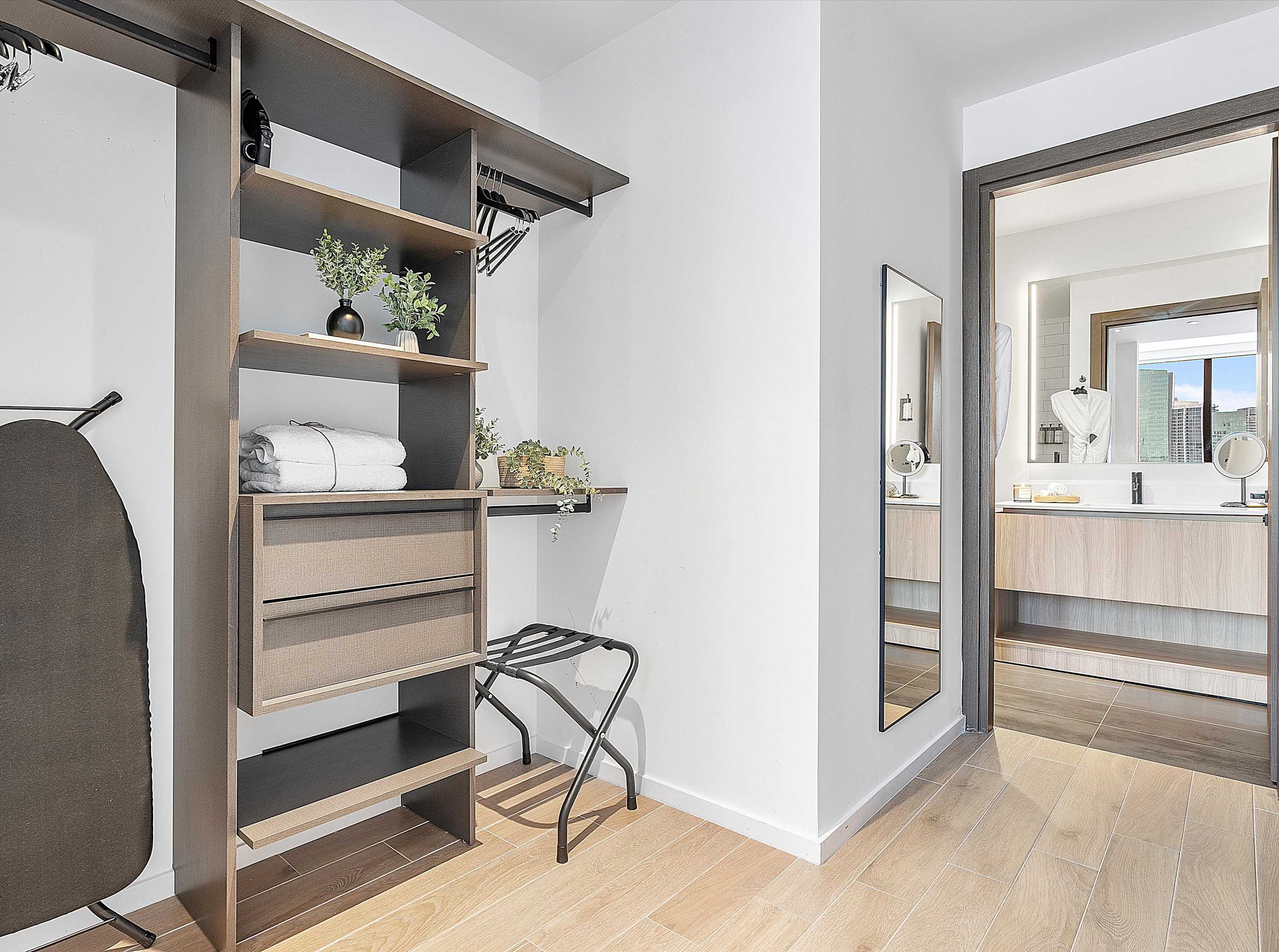 A well-organized walk-in closet or dressing area with open shelving, drawers, and hanging space in dark wood, featuring an ironing board, neatly folded towels, and a luggage rack, with light wood-look flooring.