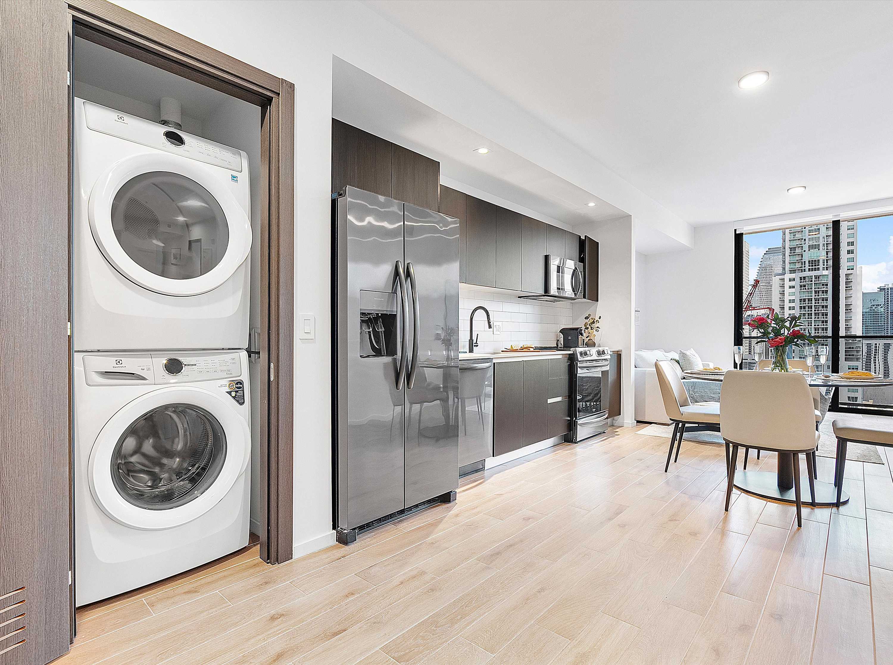 A sleek, open-concept kitchen featuring dark wood cabinetry, stainless steel appliances, a white subway tile backsplash, and light wood-look flooring, with a stacked washer and dryer unit neatly tucked into an adjacent closet.