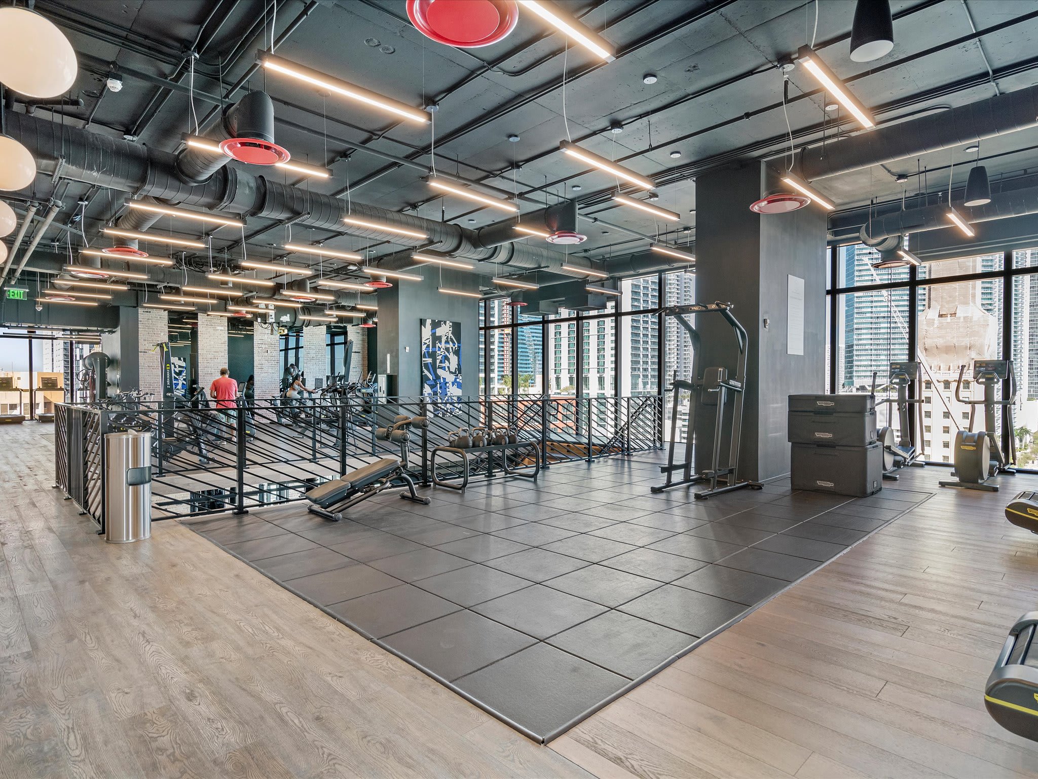 A spacious and well-appointed fitness center featuring various weight machines and free weights, designed with a multi-level layout, industrial-style ceilings, and large windows providing dynamic city views.