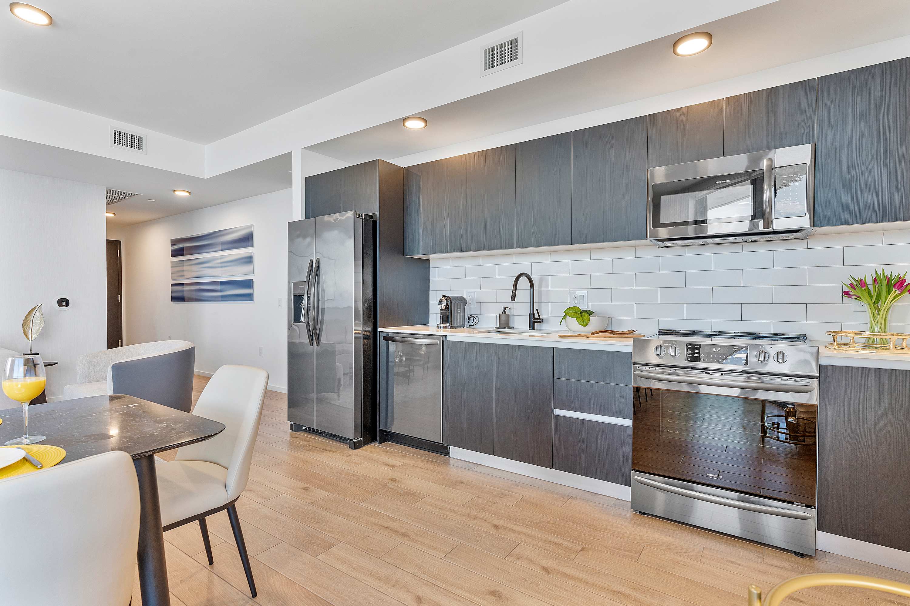 Contemporary kitchen featuring dark gray flat-panel cabinetry, white subway tile backsplash, light countertops, and stainless steel appliances including a refrigerator, dishwasher, oven, and microwave.