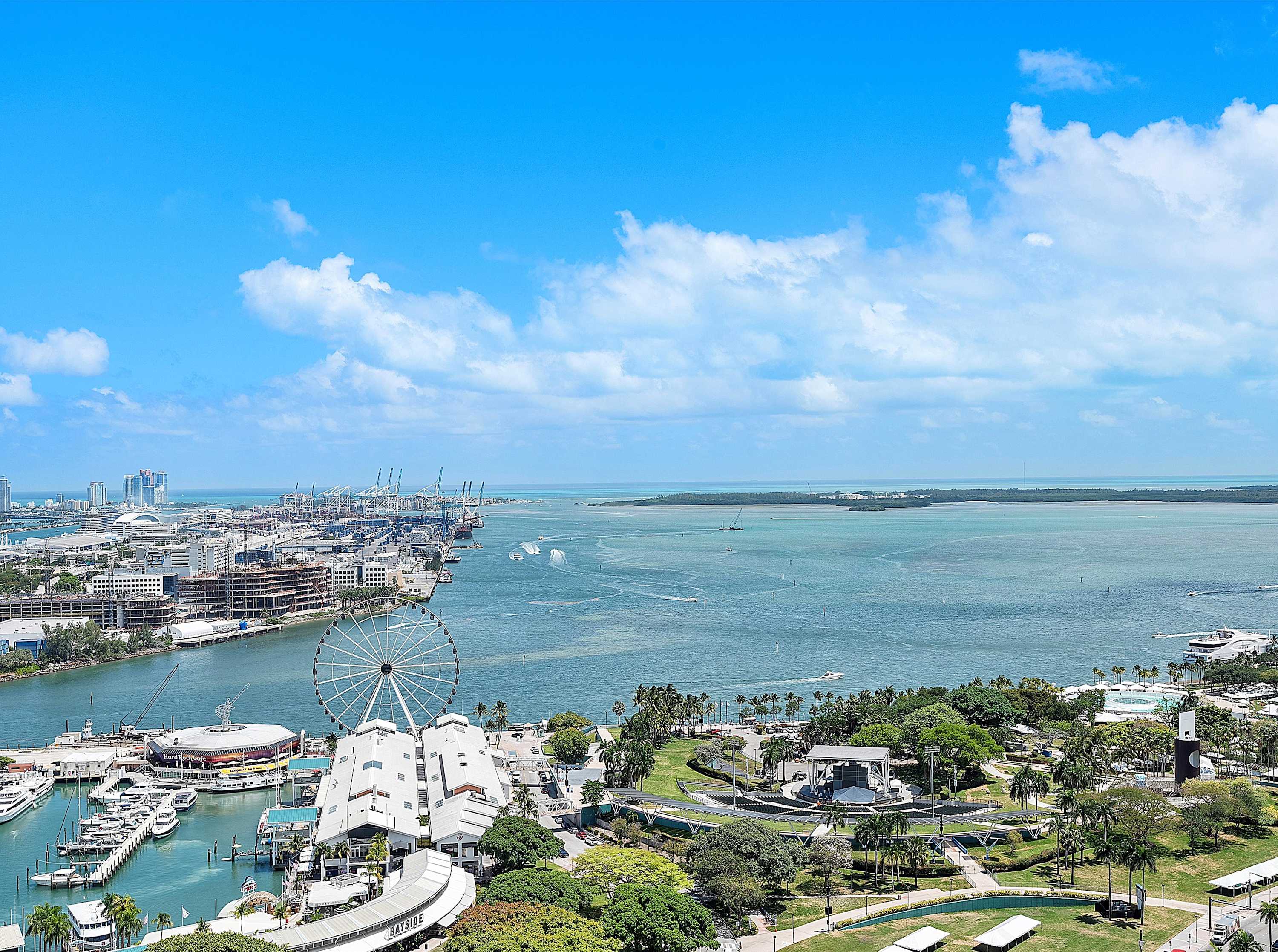A panoramic view of a dynamic waterfront city, featuring a large bay dotted with boats, a prominent Ferris wheel, lush green parks, and numerous urban structures under a brilliant blue sky.