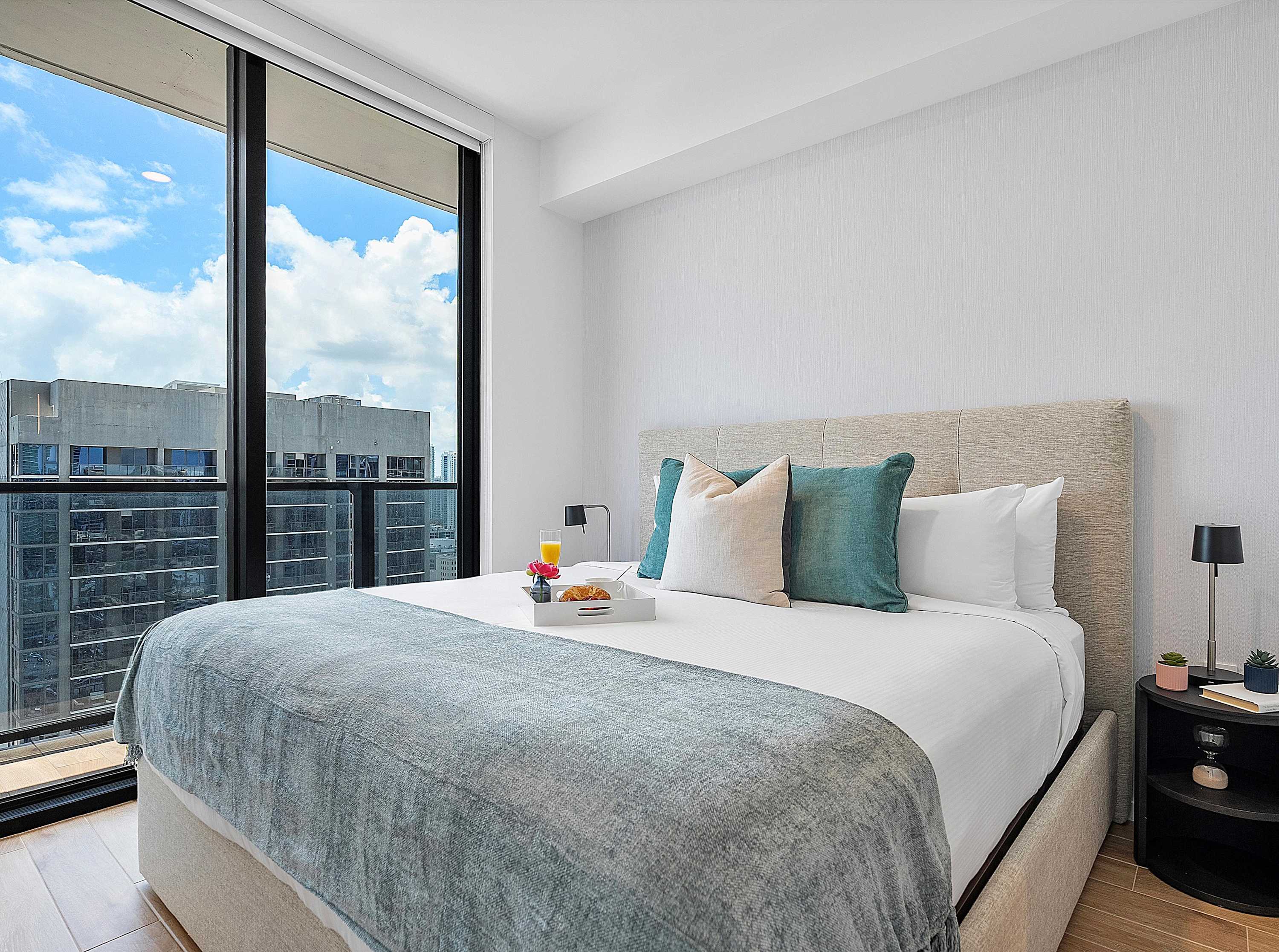 Serene bedroom featuring a light upholstered king-size bed with white and teal bedding, flanked by nightstands, and offering city views through expansive floor-to-ceiling windows.