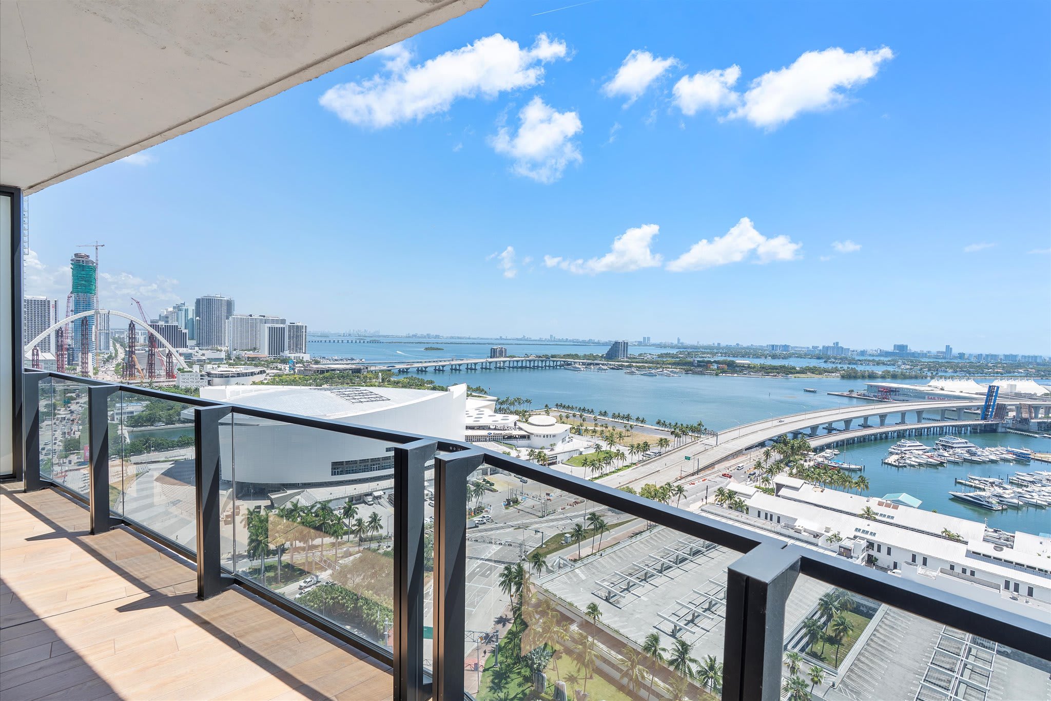 A wide-angle view from a high-rise balcony with a clear glass railing, showcasing a sprawling cityscape with modern buildings, multiple bridges, and a bustling marina under a bright blue sky with scattered clouds.