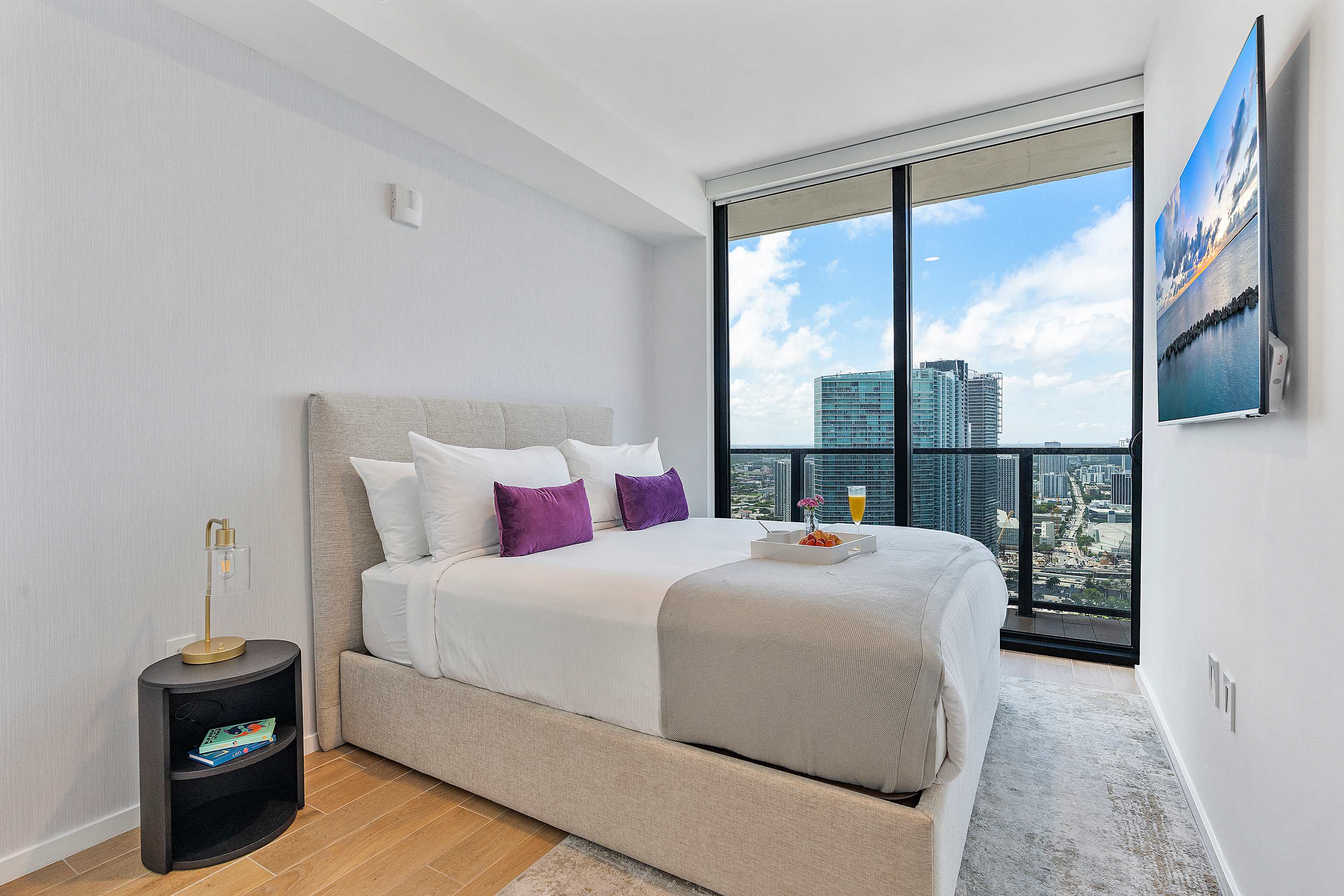 Bright bedroom with a light upholstered king-size bed, white bedding, and purple accent pillows, featuring floor-to-ceiling windows that offer expansive city and water views.