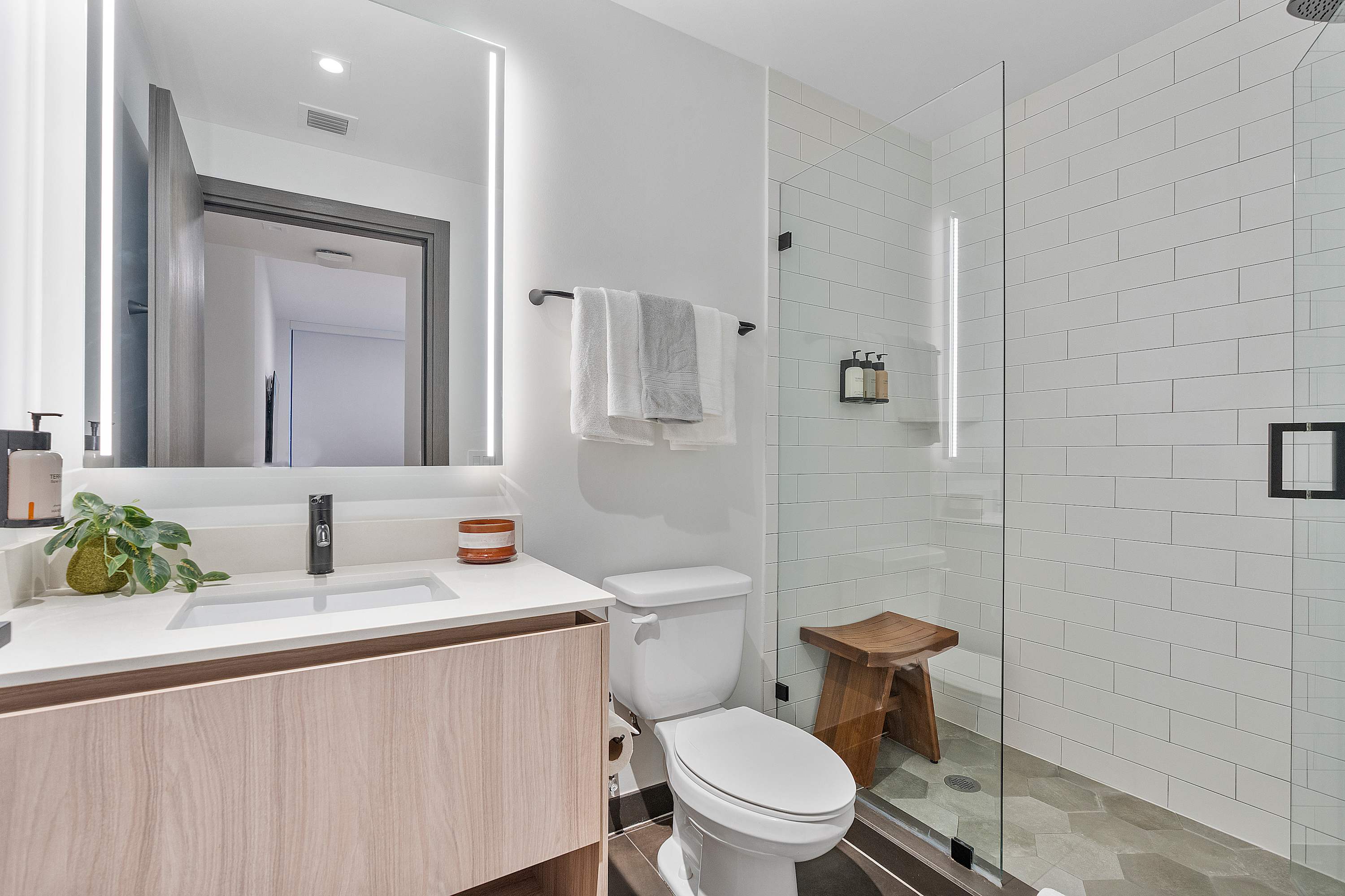 Modern bathroom with a light wood vanity, white countertop, illuminated mirror, and a walk-in shower with white subway tiles and a glass enclosure, featuring a wooden stool.