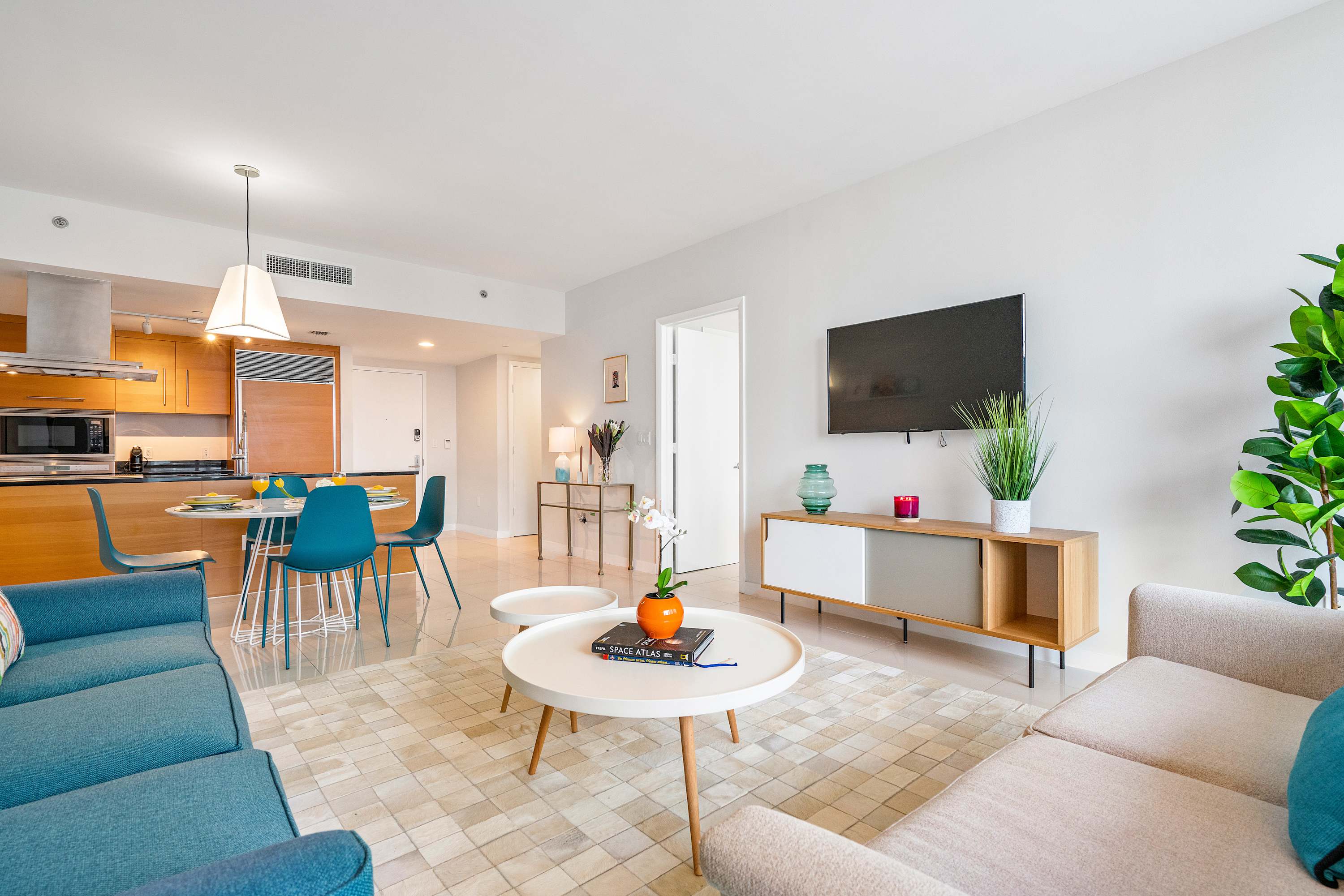 Modern open-concept living area flowing into a dining space and kitchen, featuring a teal sofa, a beige sofa, a wooden media console, and a white dining table with teal chairs, all on white tiled floors.