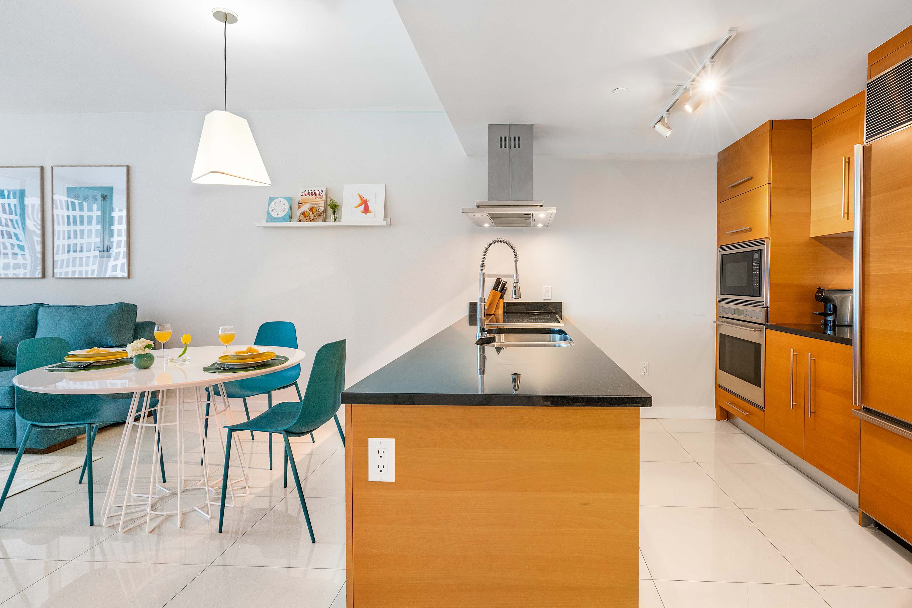 Open-concept kitchen and dining area with a large wooden island featuring a sink, stainless steel appliances, and light wood cabinetry. A round white dining table with teal chairs is visible.