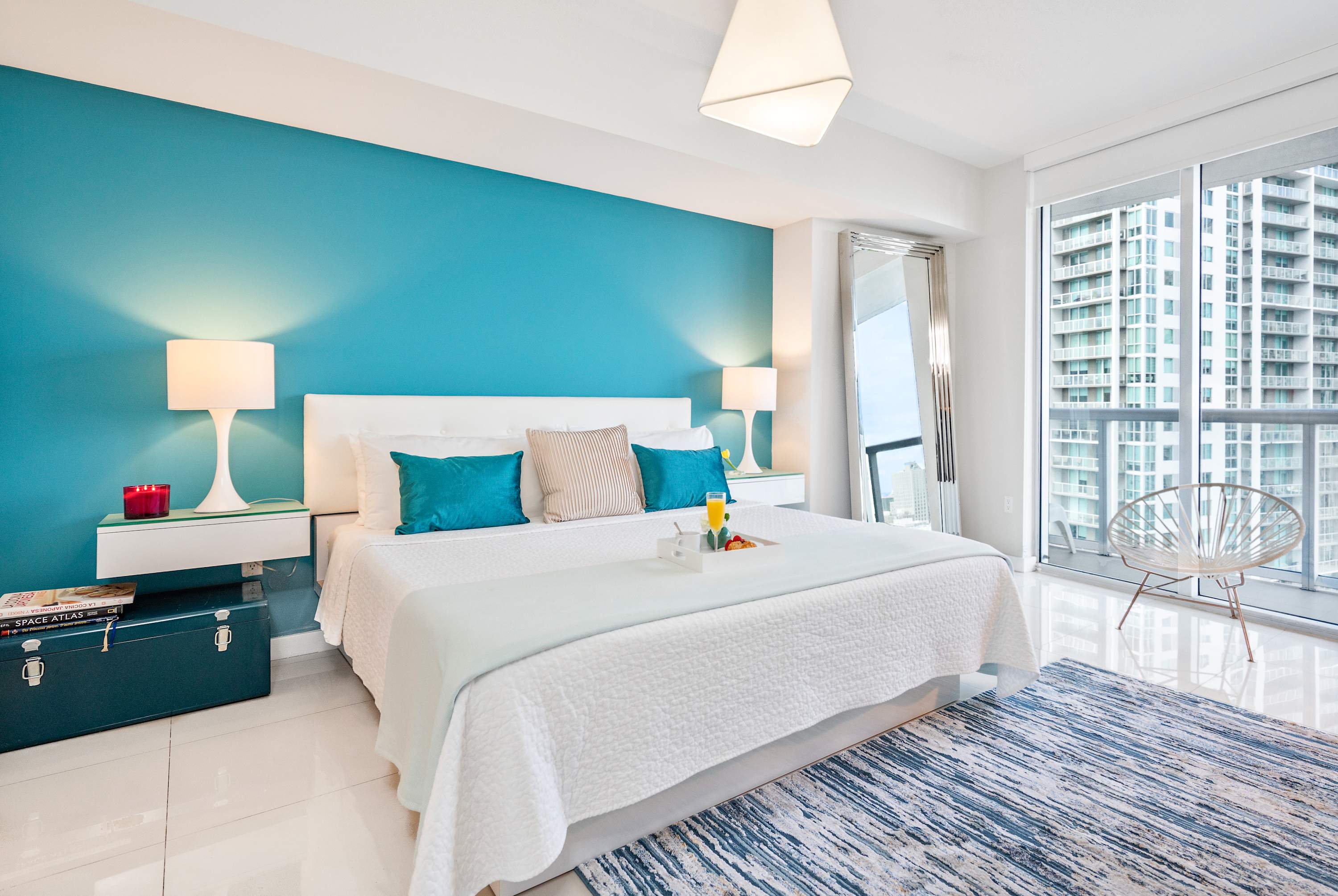 Bright bedroom with a white bed, a vibrant teal accent wall behind the headboard, white nightstands with lamps, and large sliding glass doors leading to a balcony with city views.