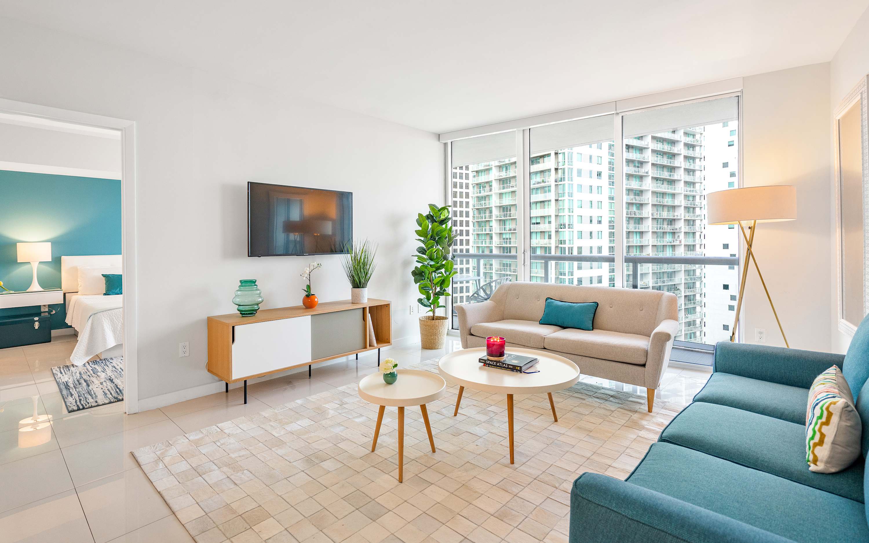 Open-plan living room with a teal sofa and a beige sofa, a wooden media console with a TV, and two round coffee tables, offering a view into the bedroom through an open doorway.