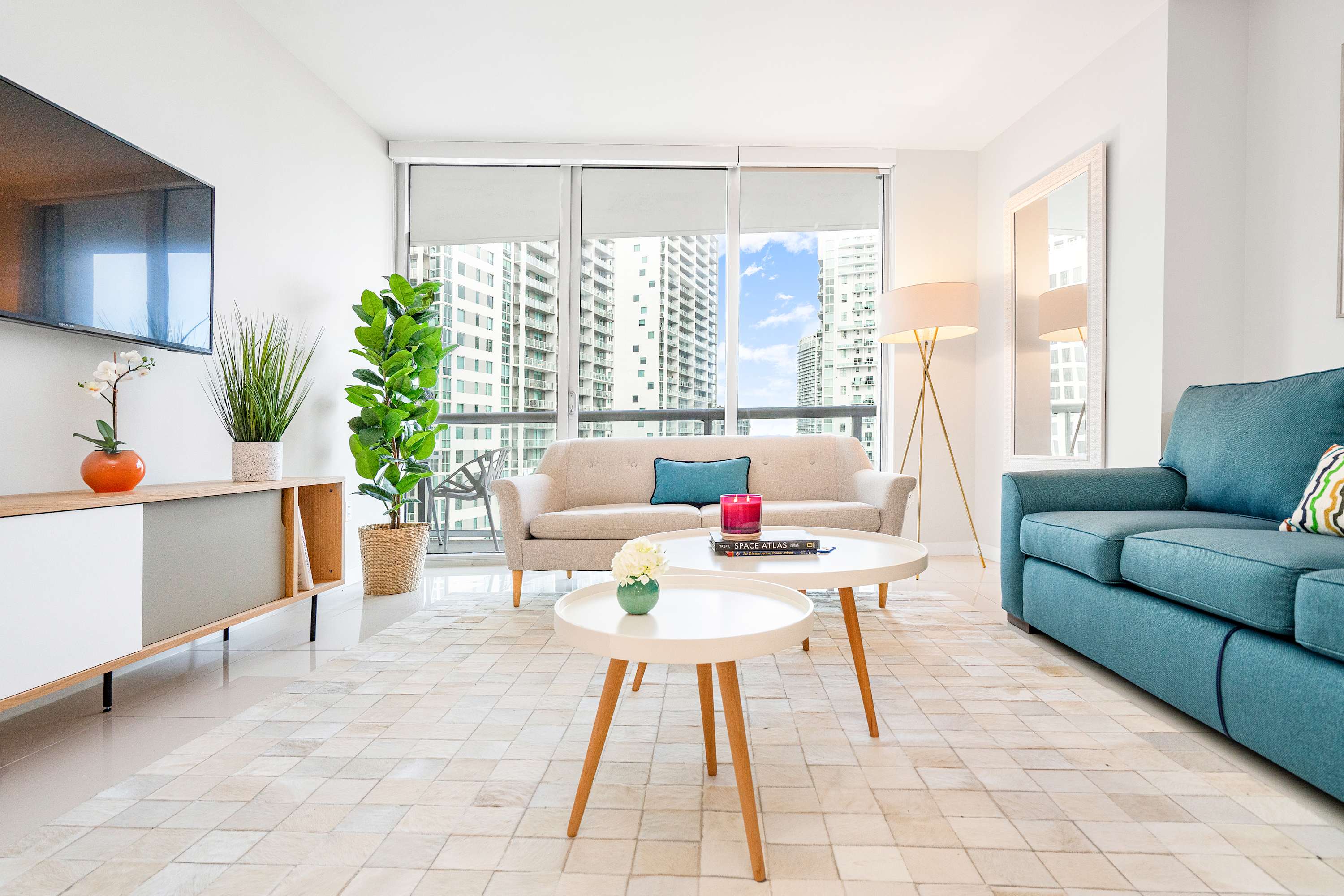 Contemporary living room with white tiled floors, a teal sofa, a beige sofa, a wooden media console with a TV, and two round white coffee tables, all illuminated by natural light from floor-to-ceiling windows with city views.