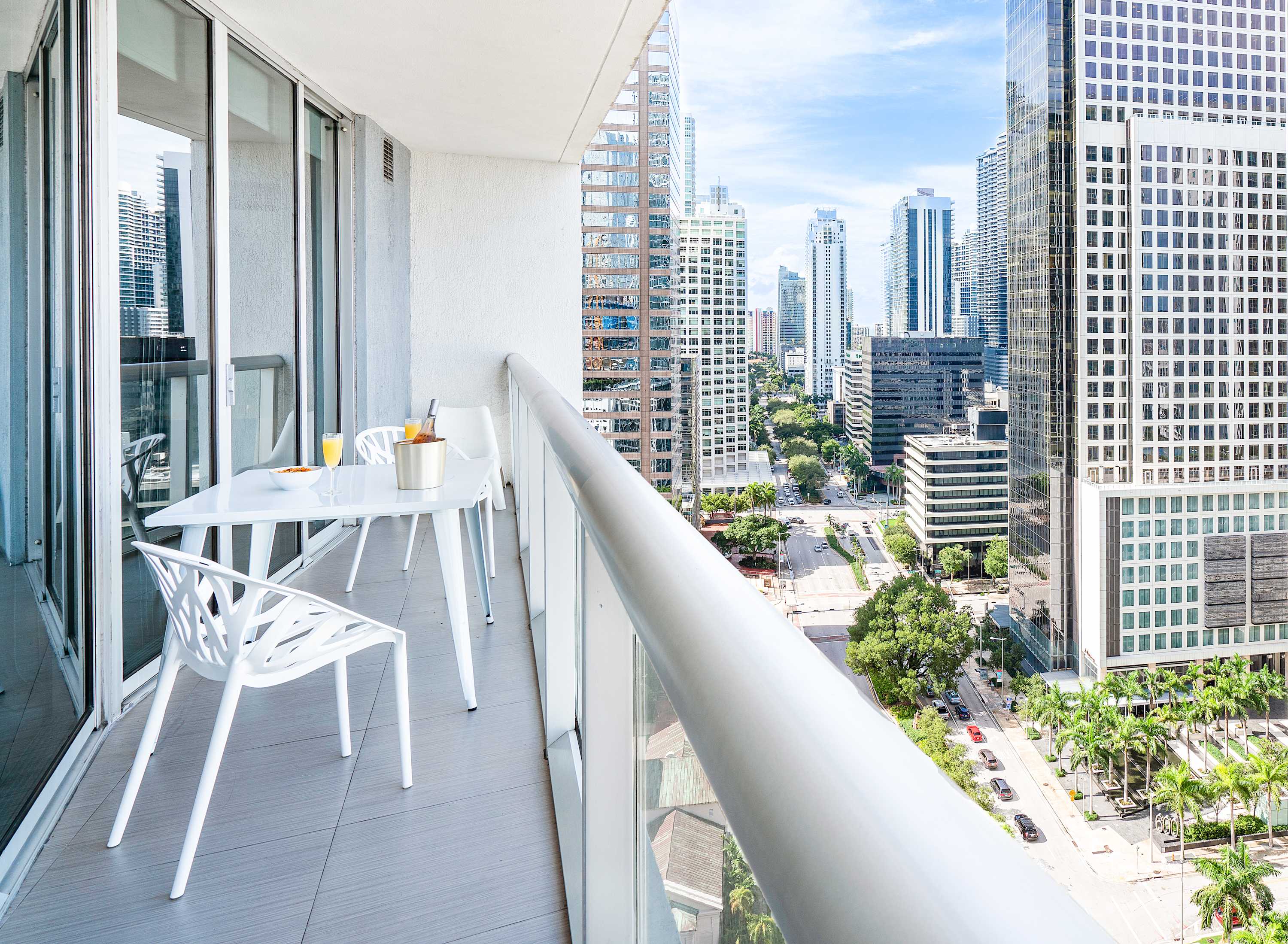 Spacious balcony with white outdoor furniture, including a dining table and chairs, offering panoramic views of a bustling city skyline with numerous high-rise buildings and a street below.