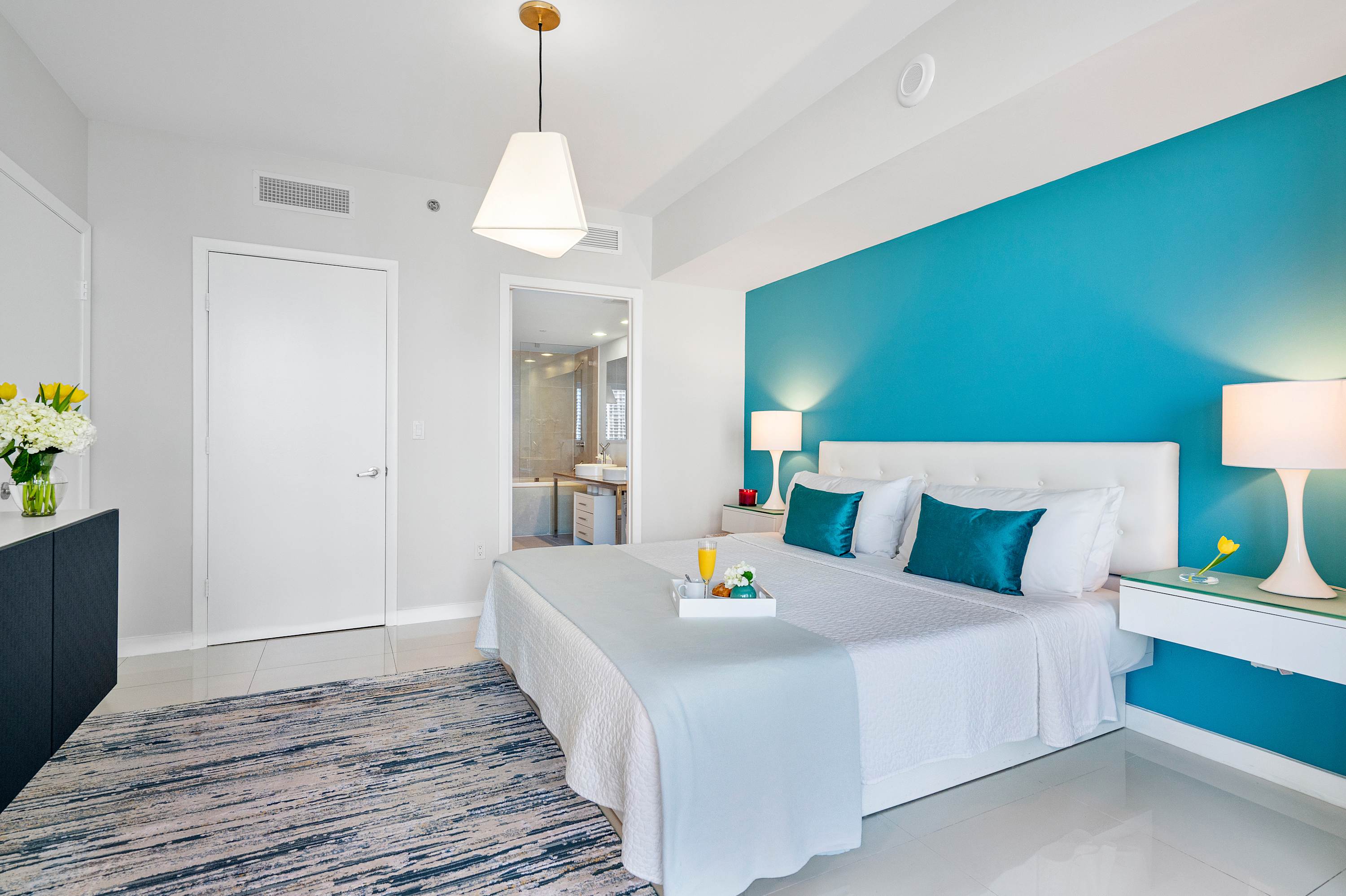 Spacious bedroom featuring a white bed with teal pillows, a striking teal accent wall, white tiled floors, and a large area rug, with a glimpse of the en-suite bathroom through an open doorway.