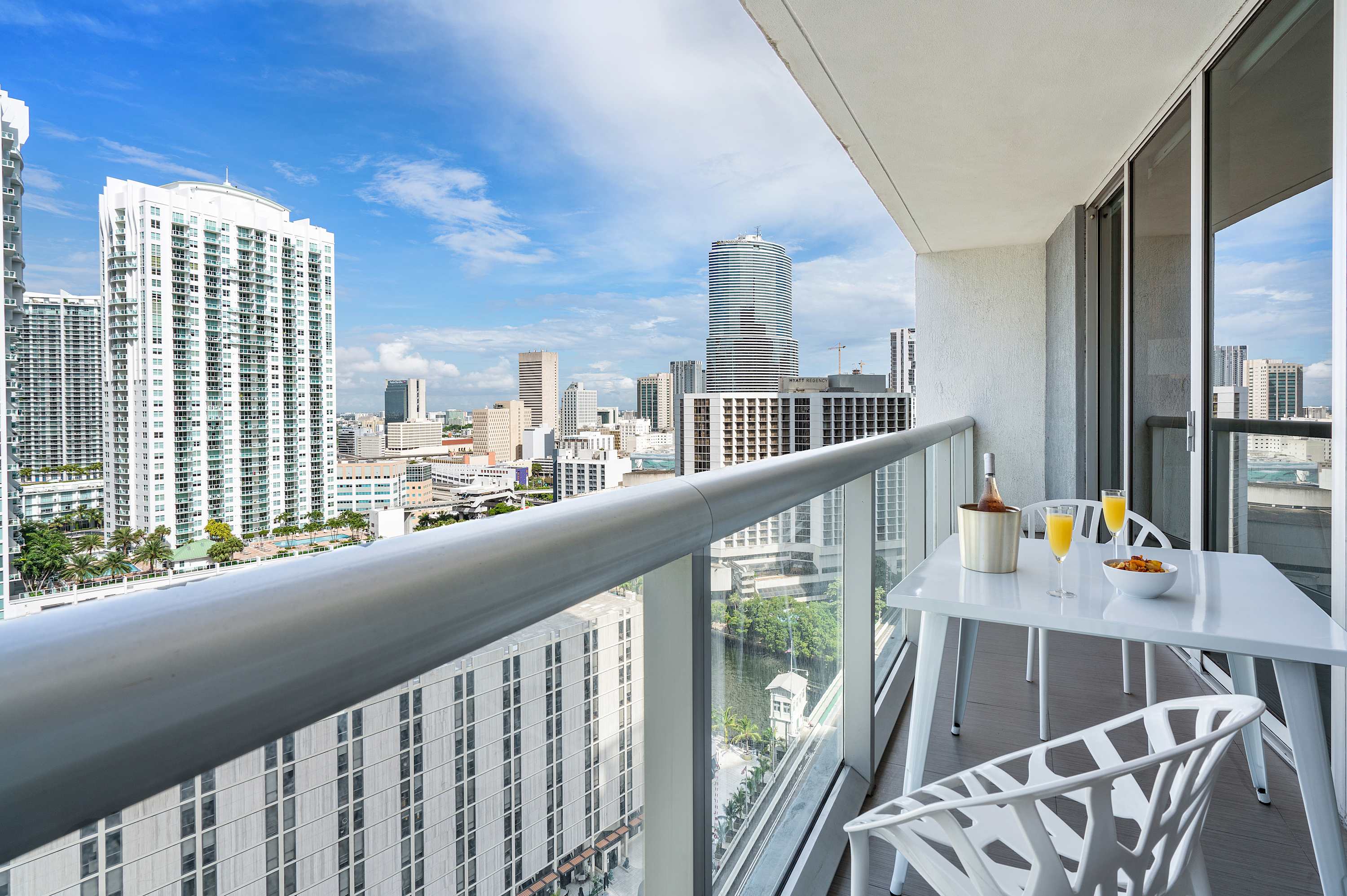 Private balcony with a white outdoor dining set, offering expansive views of the city skyline and a waterway below. The railing is modern and metallic.