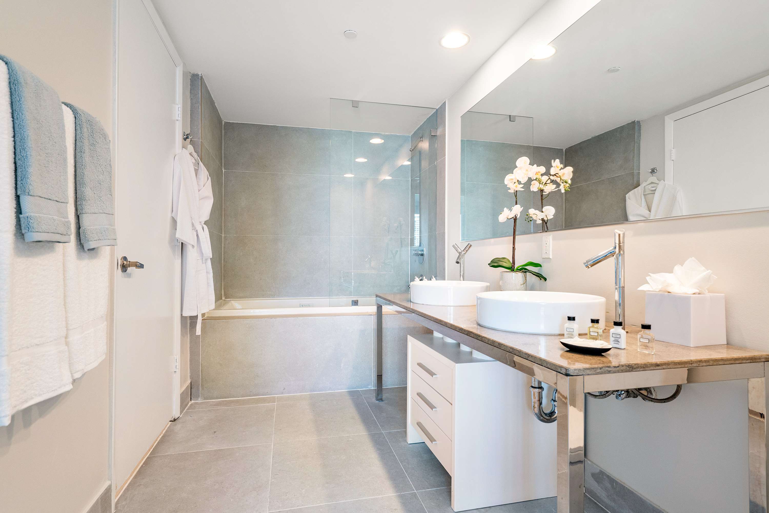 Modern bathroom featuring a long double vanity with vessel sinks, chrome faucets, a large mirror, and a built-in bathtub with light gray tiled walls. White bathrobes hang on the wall.