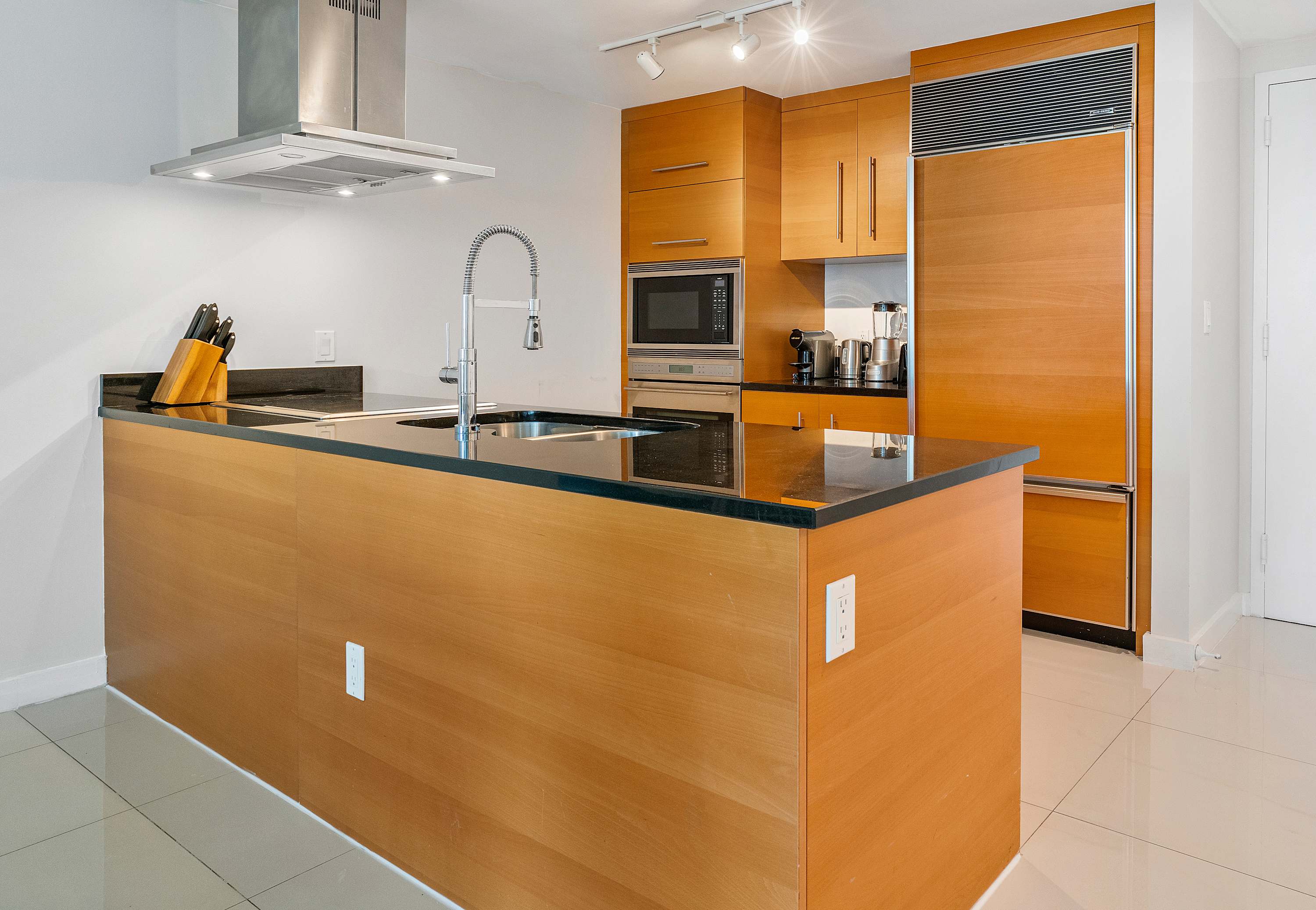 Modern kitchen featuring a large wooden island with a black countertop and integrated sink, surrounded by light wood cabinetry and stainless steel appliances including a built-in refrigerator and oven.