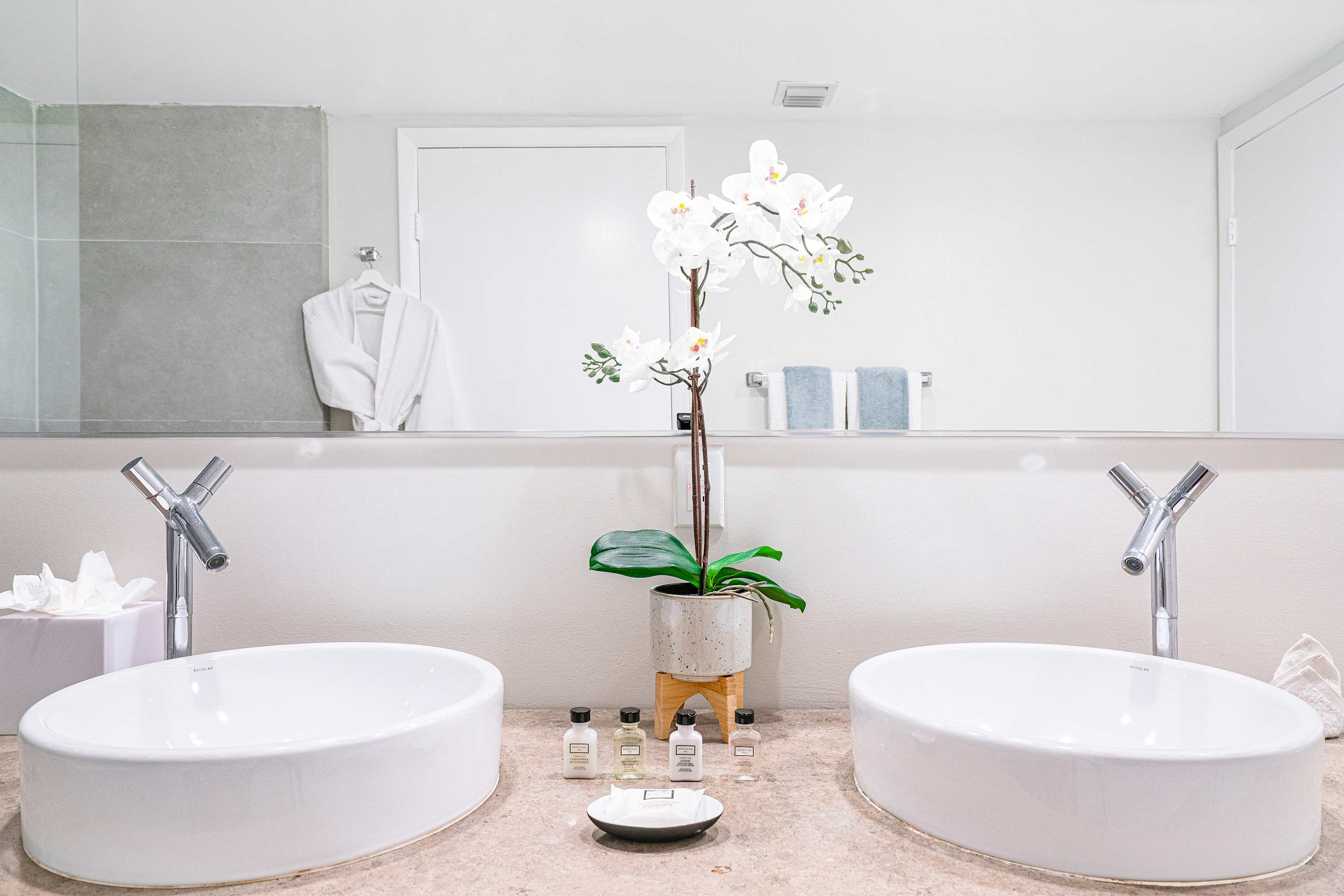Close-up of a contemporary double vanity with white vessel sinks, sleek chrome faucets, and a large mirror, adorned with a white orchid plant and small toiletries.